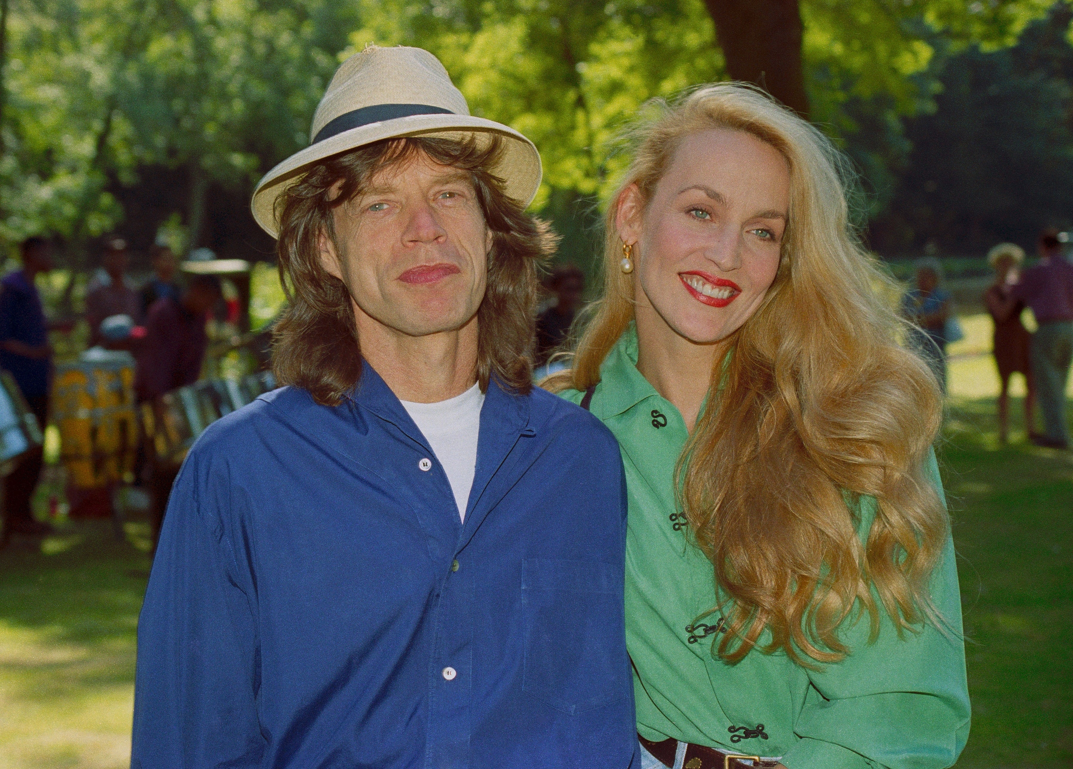 Two people outdoors; one wears a hat and casual shirt, the other has long hair and a green blouse. They are smiling and standing closely