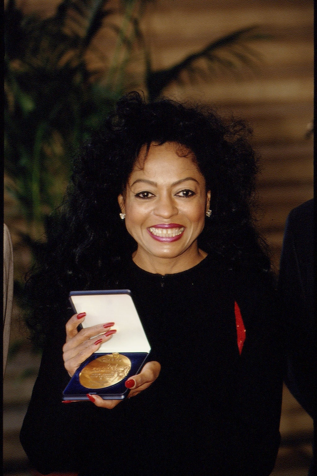 Person smiling, holding a medal in an open case. They're wearing elegant attire with wavy hair. Plants are in the background