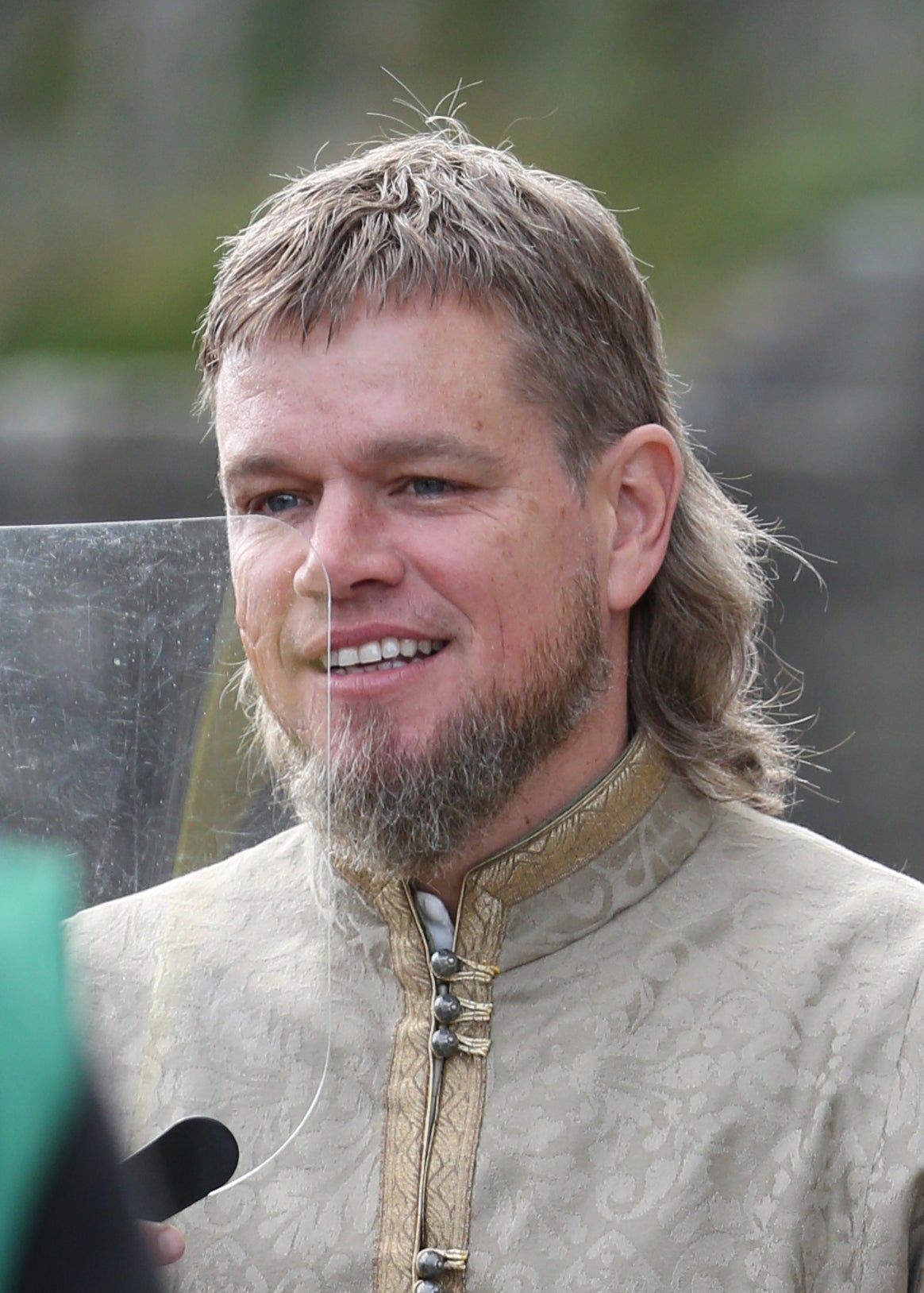 A person in ornate medieval-style costume, smiling during an outdoor event, speaking with a microphone-screen in front