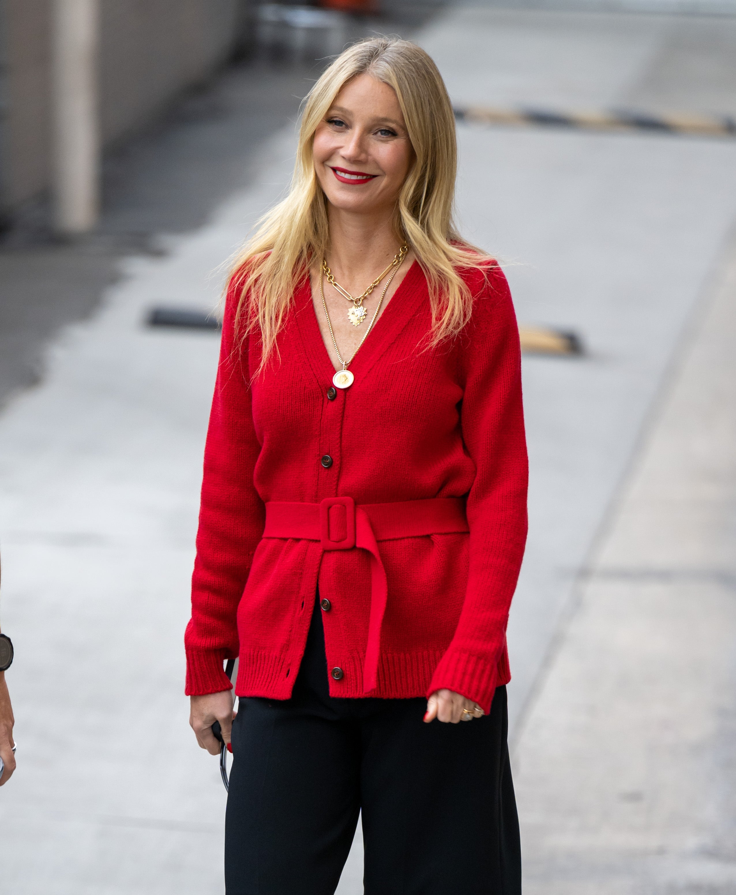 Person wearing a belted cardigan and black pants, smiling while walking outdoors