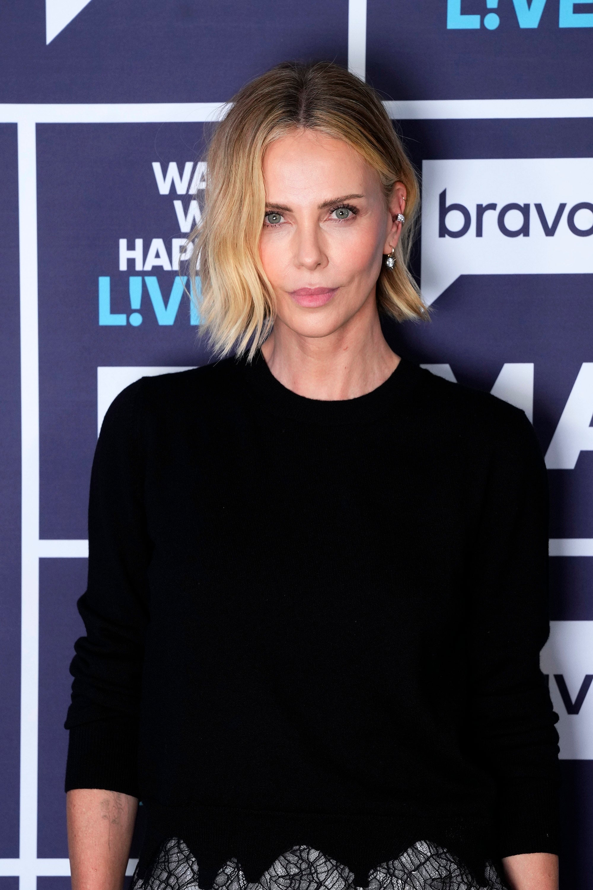 Person in a black top with short, wavy blonde hair attends a public event, standing against a backdrop with logos and text