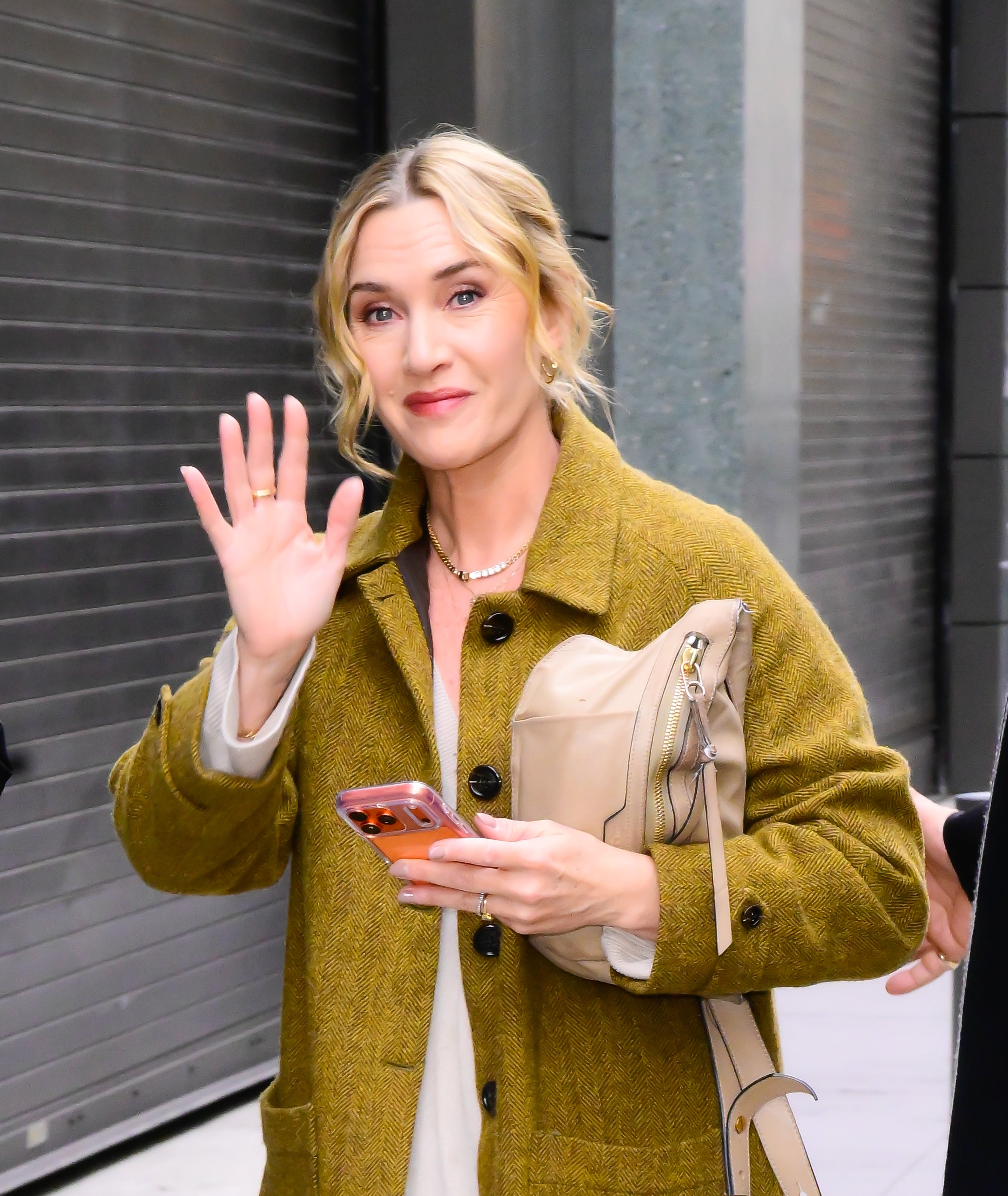 Celebrity woman waves, wearing a casual coat and holding a phone and handbag, smiling at the camera on a city street
