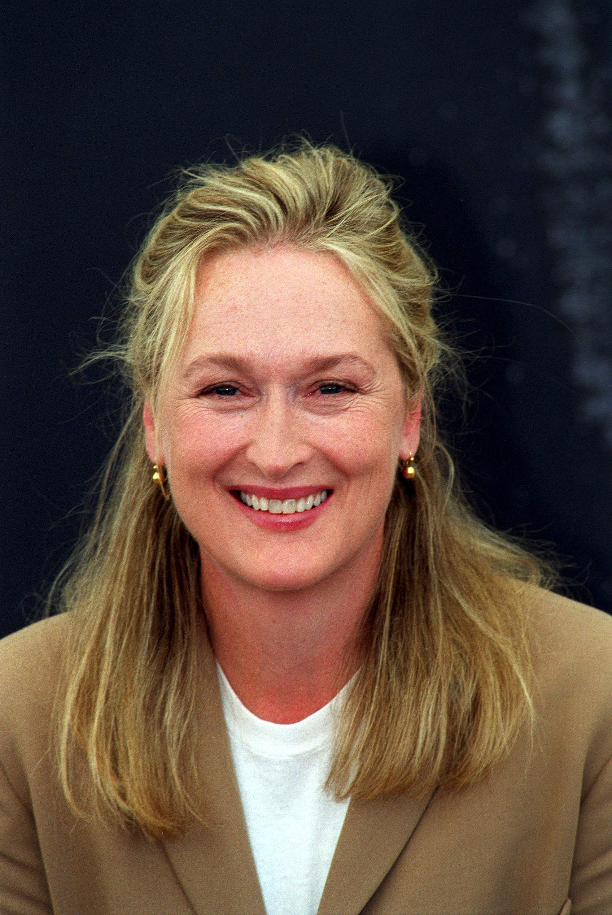 A smiling person with shoulder-length hair, wearing a blazer and a white top, poses in front of a plain backdrop