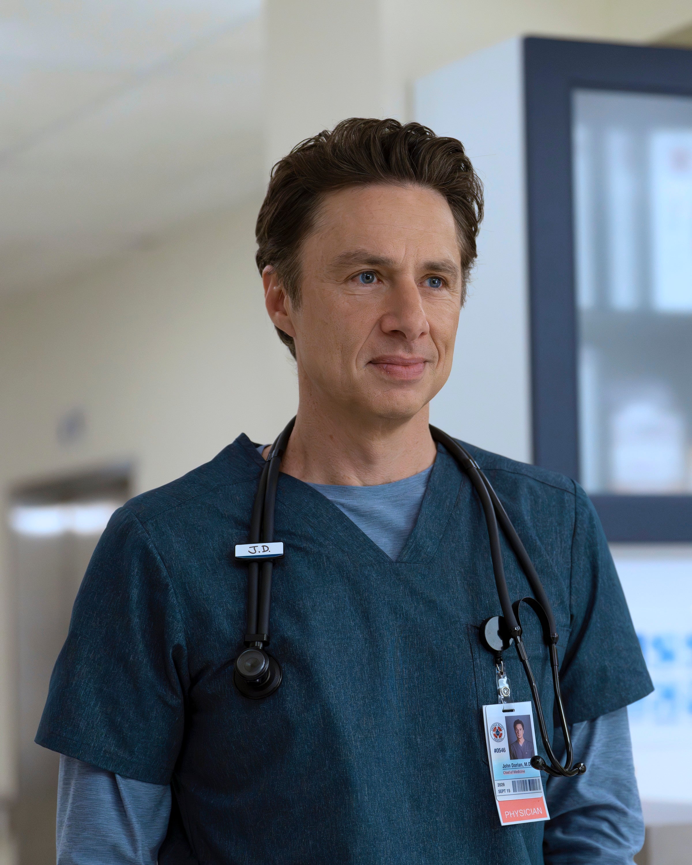 Person in a medical setting wearing scrubs with a stethoscope around their neck. They have short hair and an ID badge clipped to their clothing