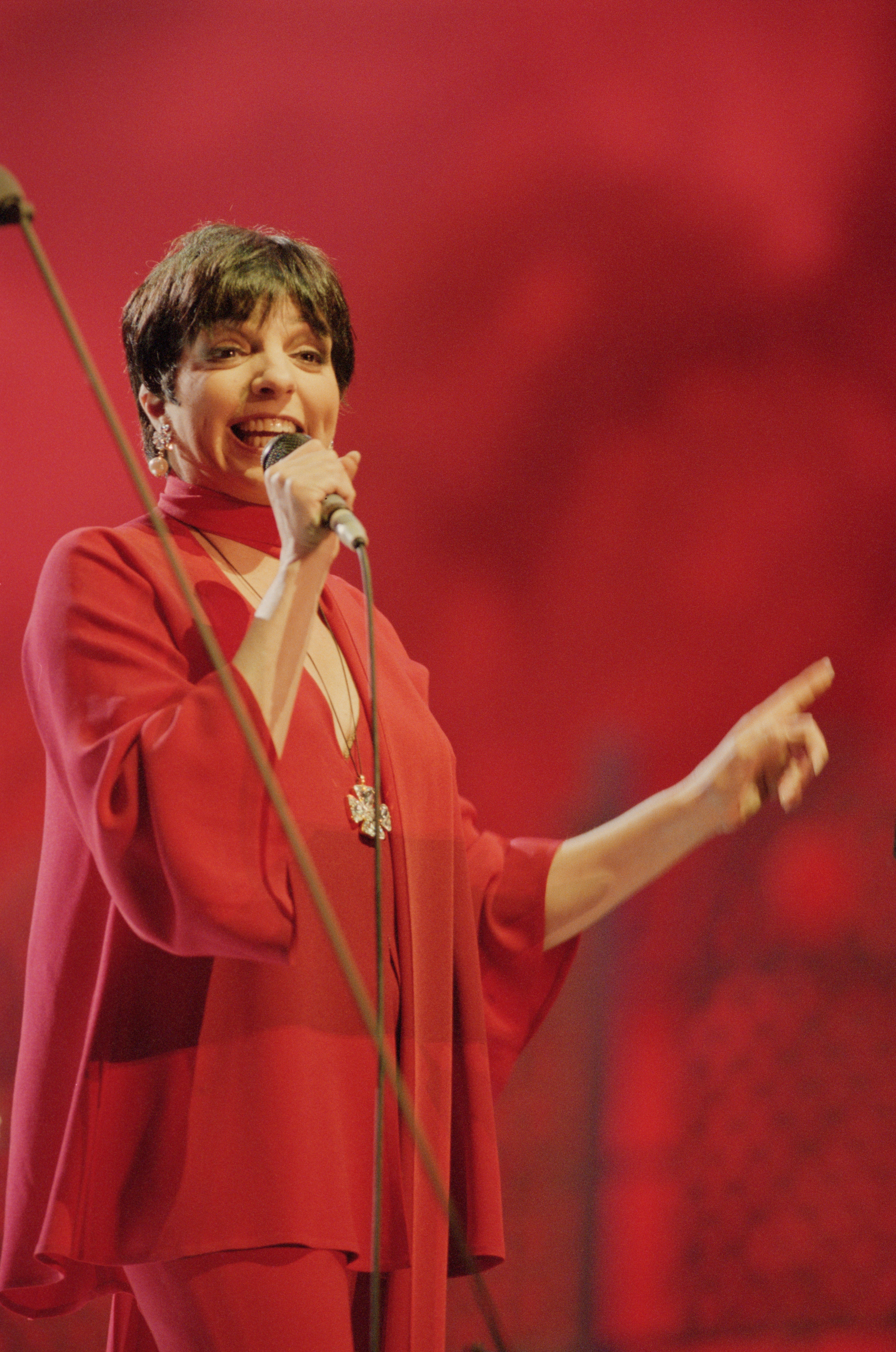 A woman in elegant, flowing attire performs on stage, holding a microphone and gesturing with her hand