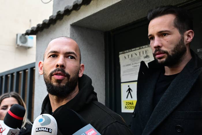 Two individuals being interviewed by media outside a building. Various microphones in the foreground