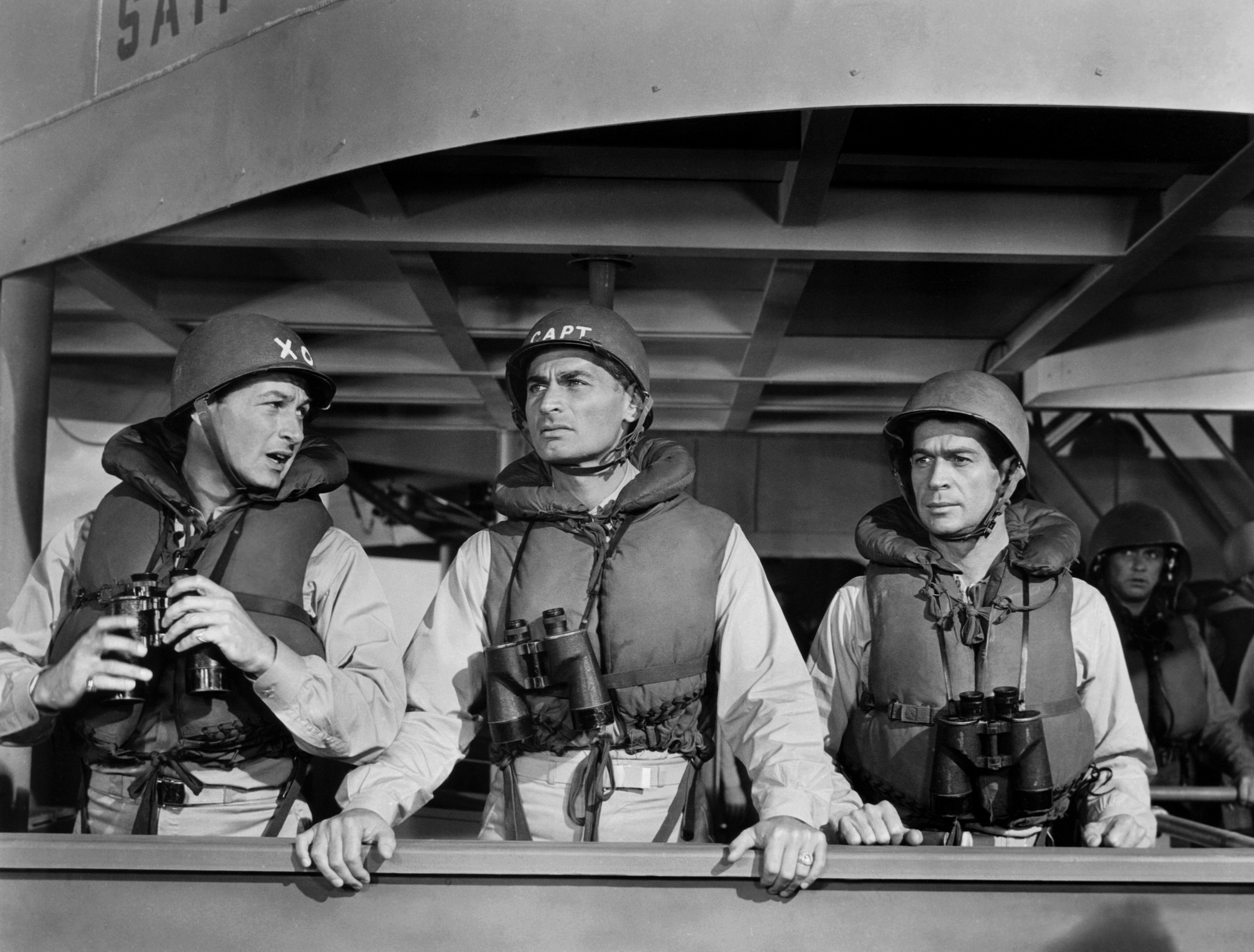 Three actors in military attire with helmets and life vests, holding binoculars, stand on a ship set in a classic film scene