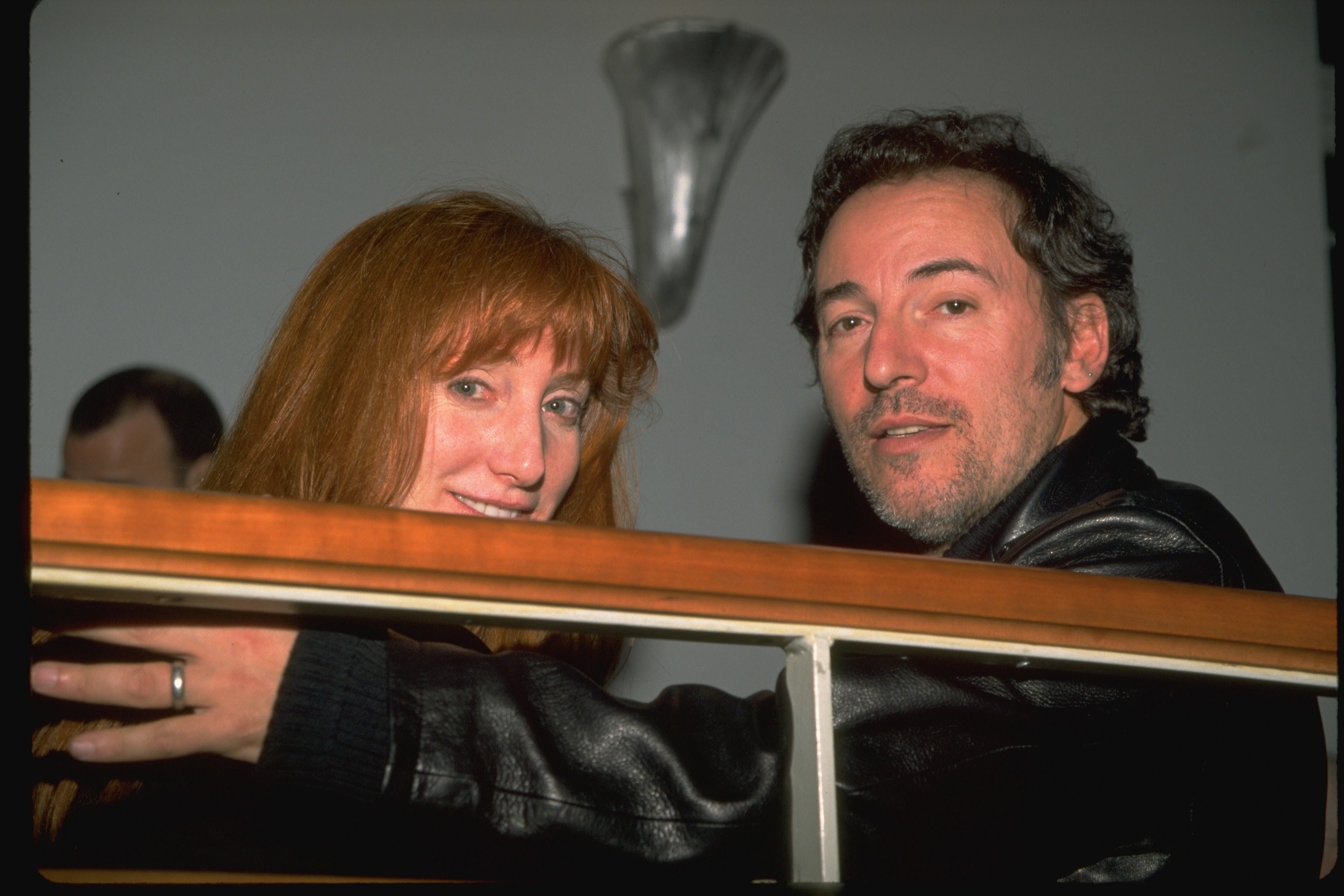 A man and woman smiling, sitting closely together, both wearing casual leather jackets