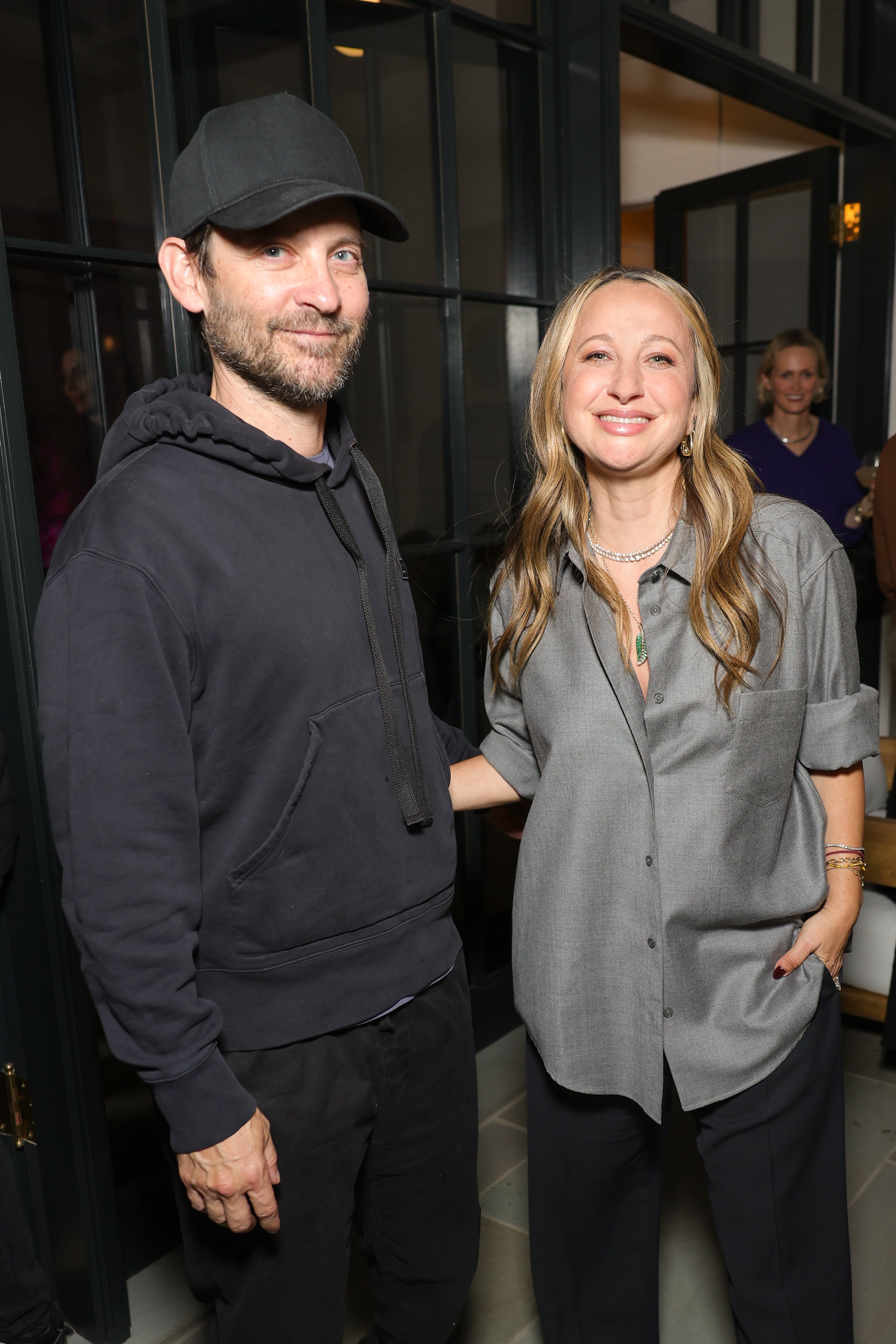 Two people stand together indoors. The person on the left wears a casual outfit with a cap and hoodie. The person on the right wears a loose-fitting shirt