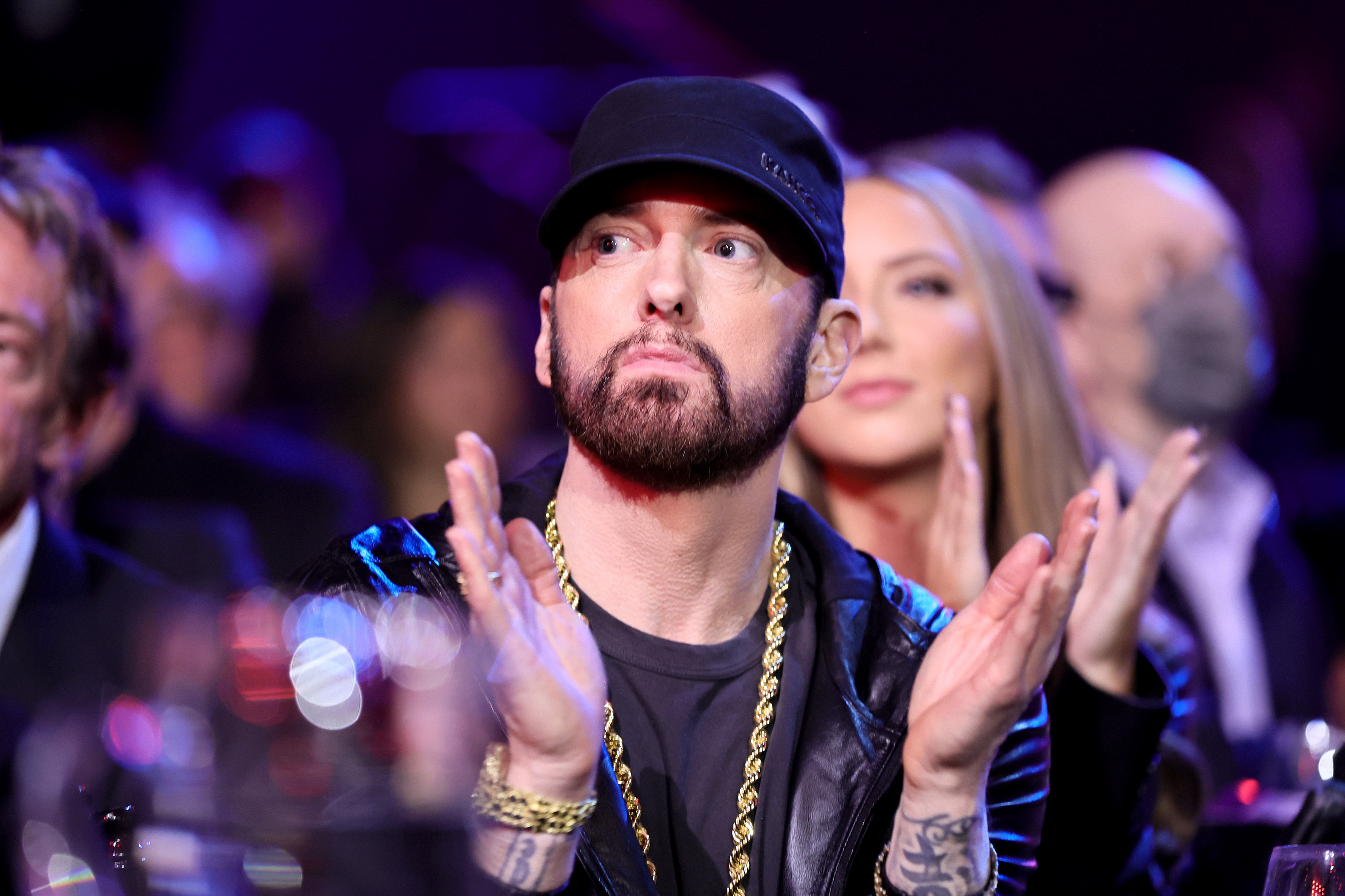 A person in a black cap and leather jacket with a gold chain is seated, clapping, and looking ahead at an event