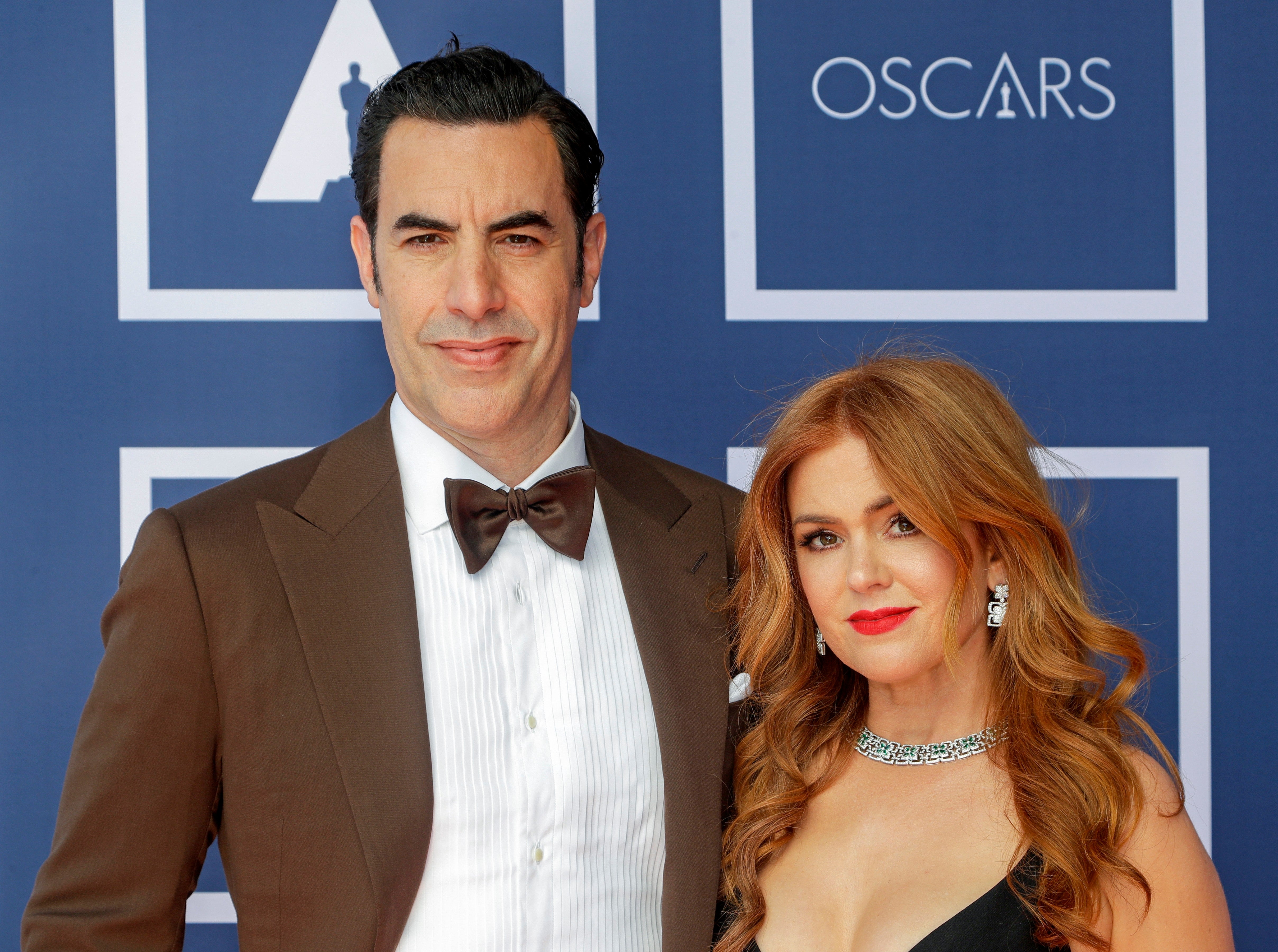 Two people on a red carpet; the man in a tuxedo and the woman in a black gown with a necklace, attending an event with an Oscars backdrop