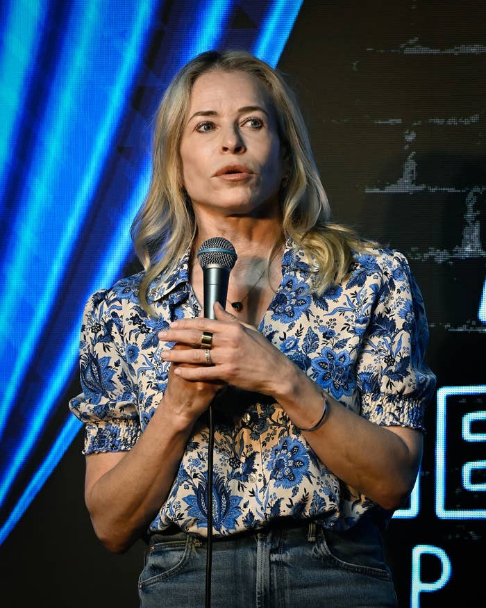 Person speaking on stage holding a microphone, wearing a patterned floral blouse and denim jeans