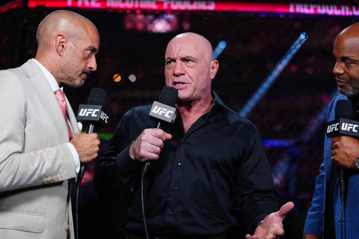 Three men, holding UFC microphones, are having a conversation in an arena. The central man gestures while speaking