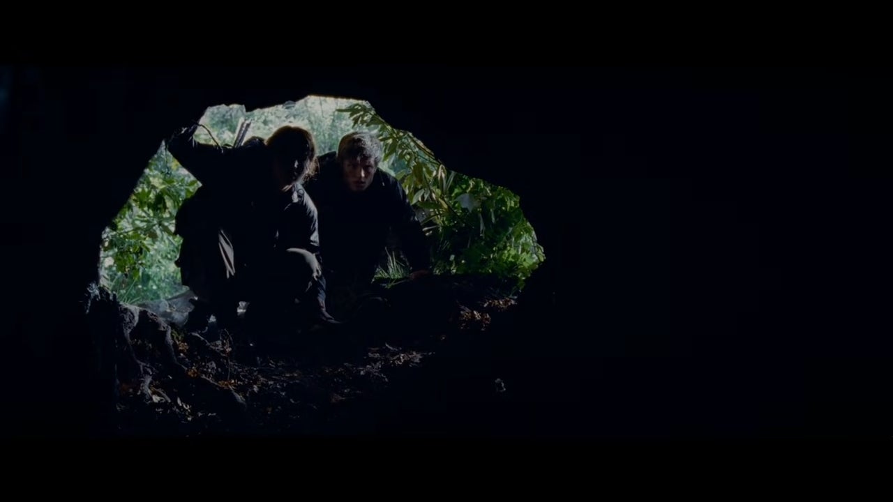 Two people crouch at the entrance of a dark cave, peering inside with cautious expressions