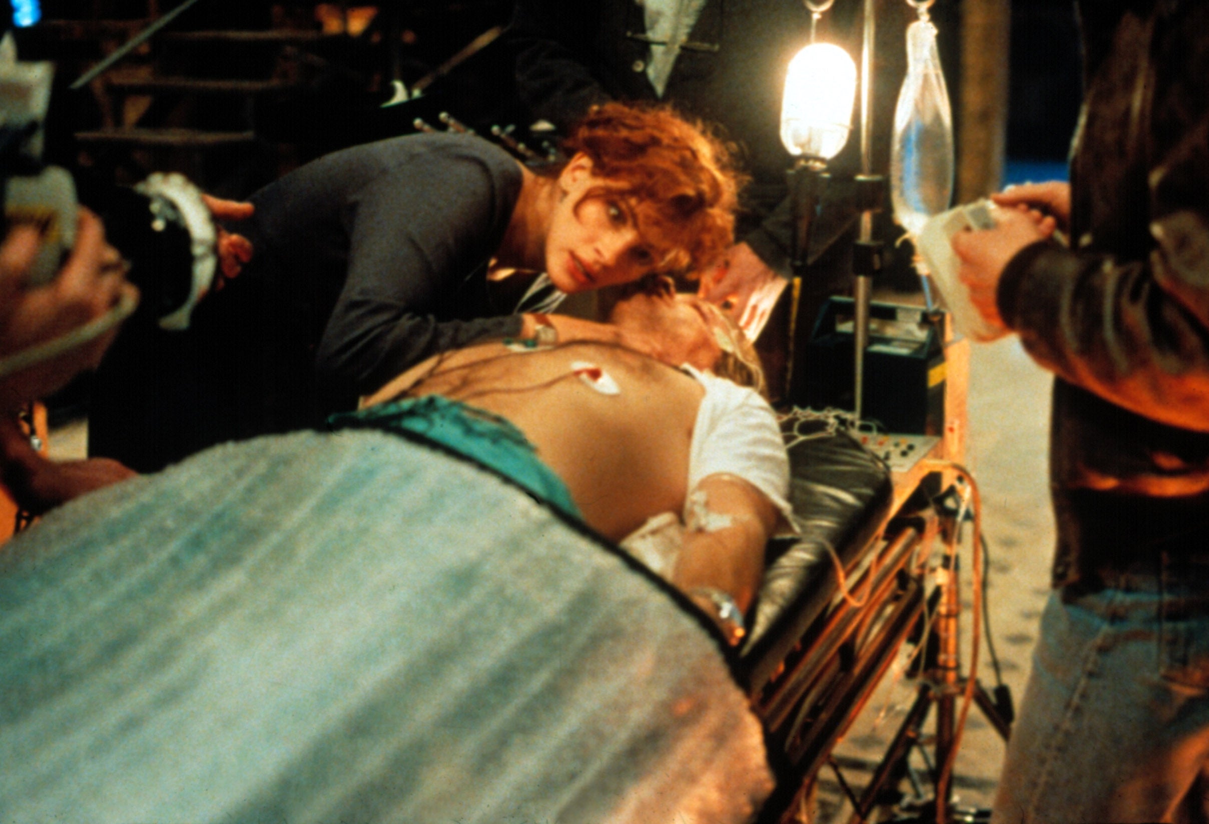 A person tends to someone on a stretcher in a dramatic scene, suggesting urgency and concern. People and equipment surround them