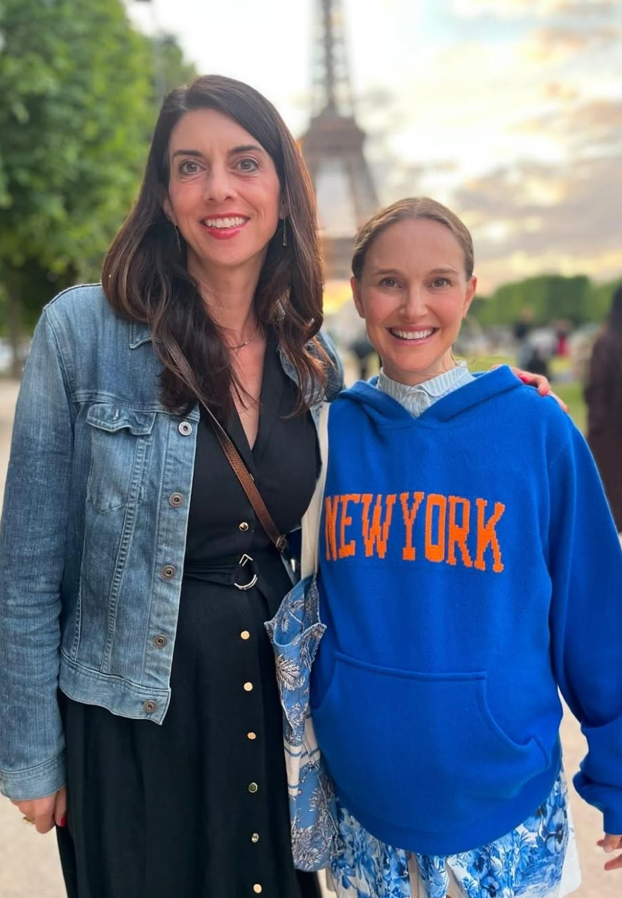 Two people smiling in front of the Eiffel Tower; one in a denim jacket and black dress, the other in a "New York" hoodie