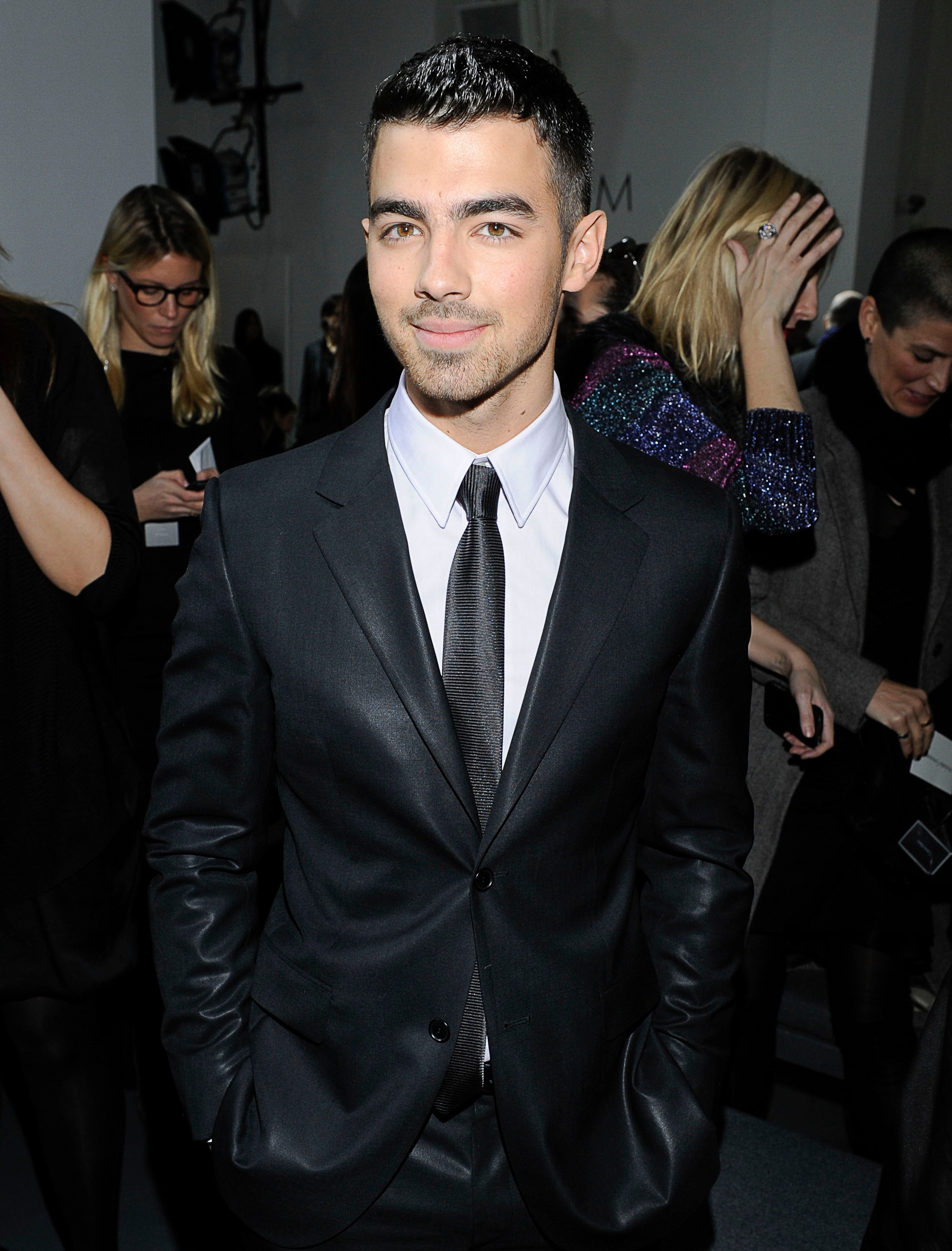 Person in a formal suit and tie at an event, hands in pockets. People in background, some interacting with the event displays