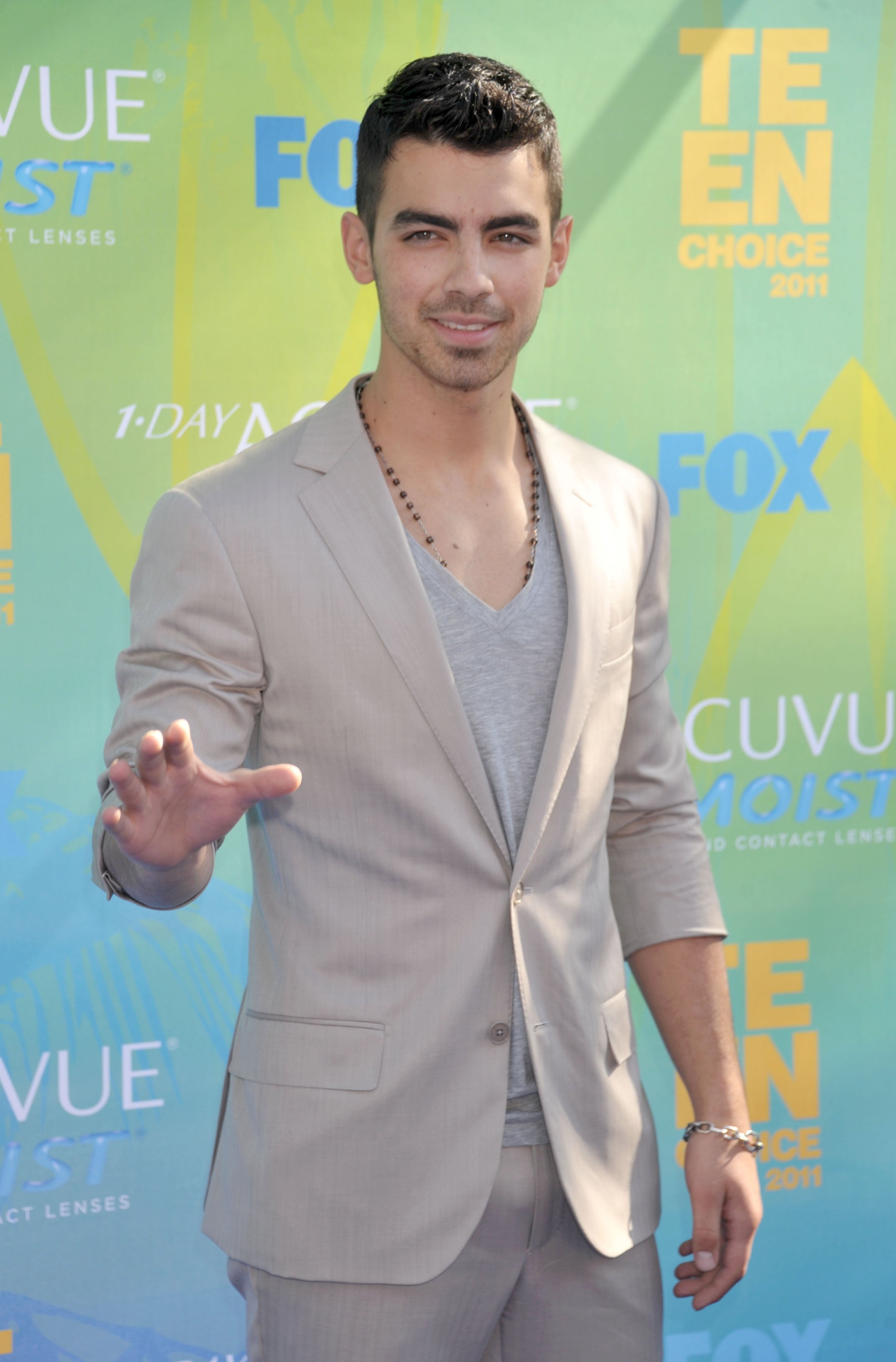 Person in a light suit with V-neck shirt poses on a red carpet event with sponsor logos in the background