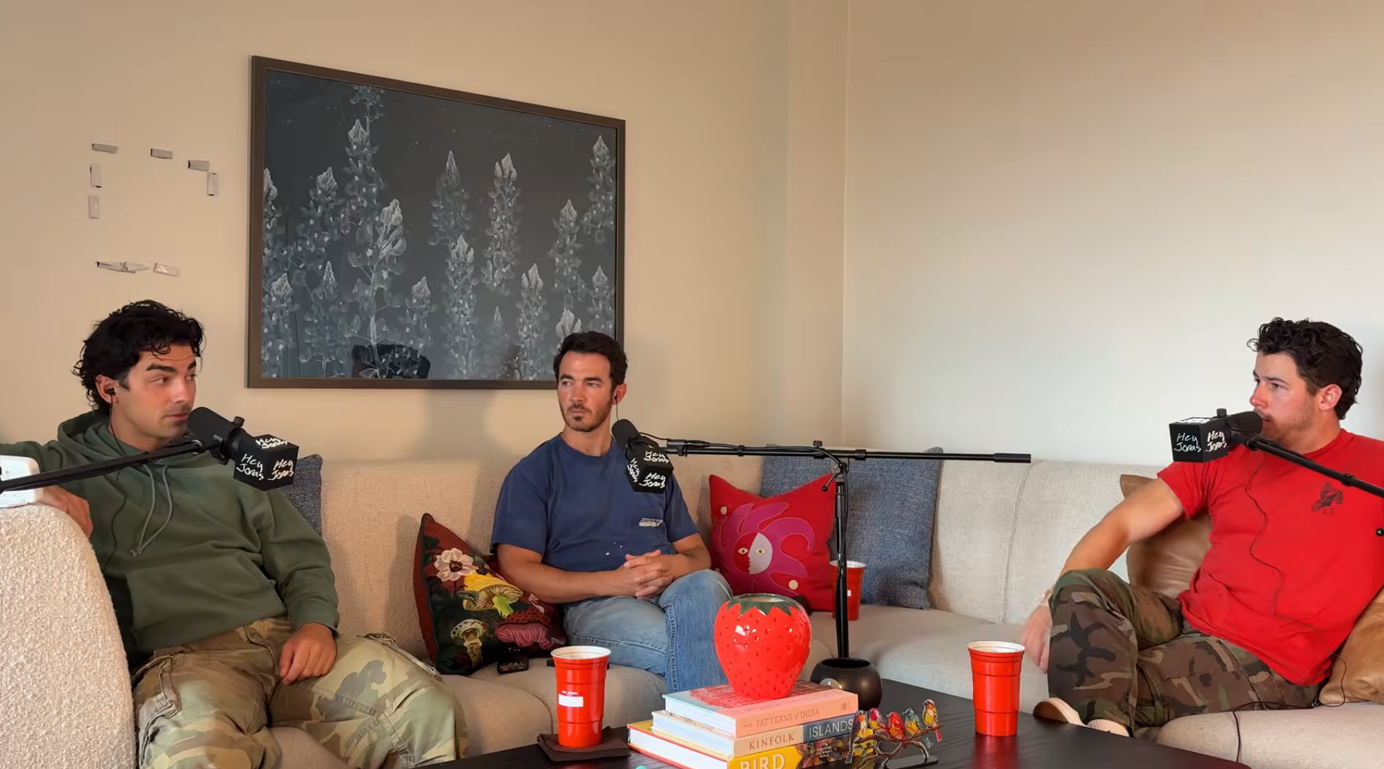 Three people sit on a cozy couch with microphones, engaged in a podcast discussion. They are casually dressed, and the setting is informal