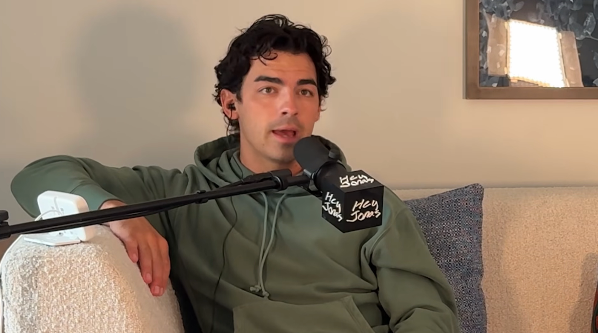 Person sitting on a couch, speaking into a microphone, wearing a hoodie with headphones on
