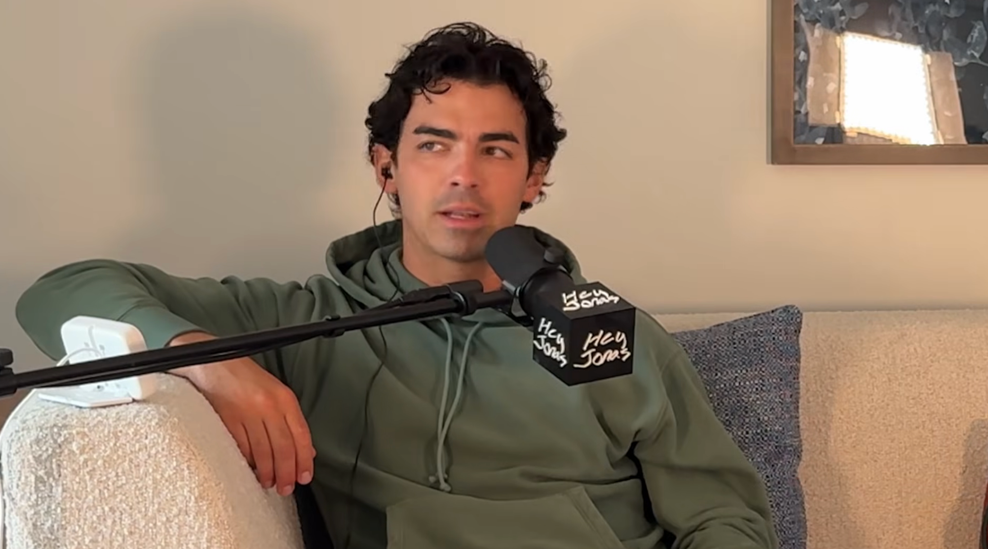Person wearing a hoodie sits on a couch, speaking into a microphone labeled "Hey Joe."