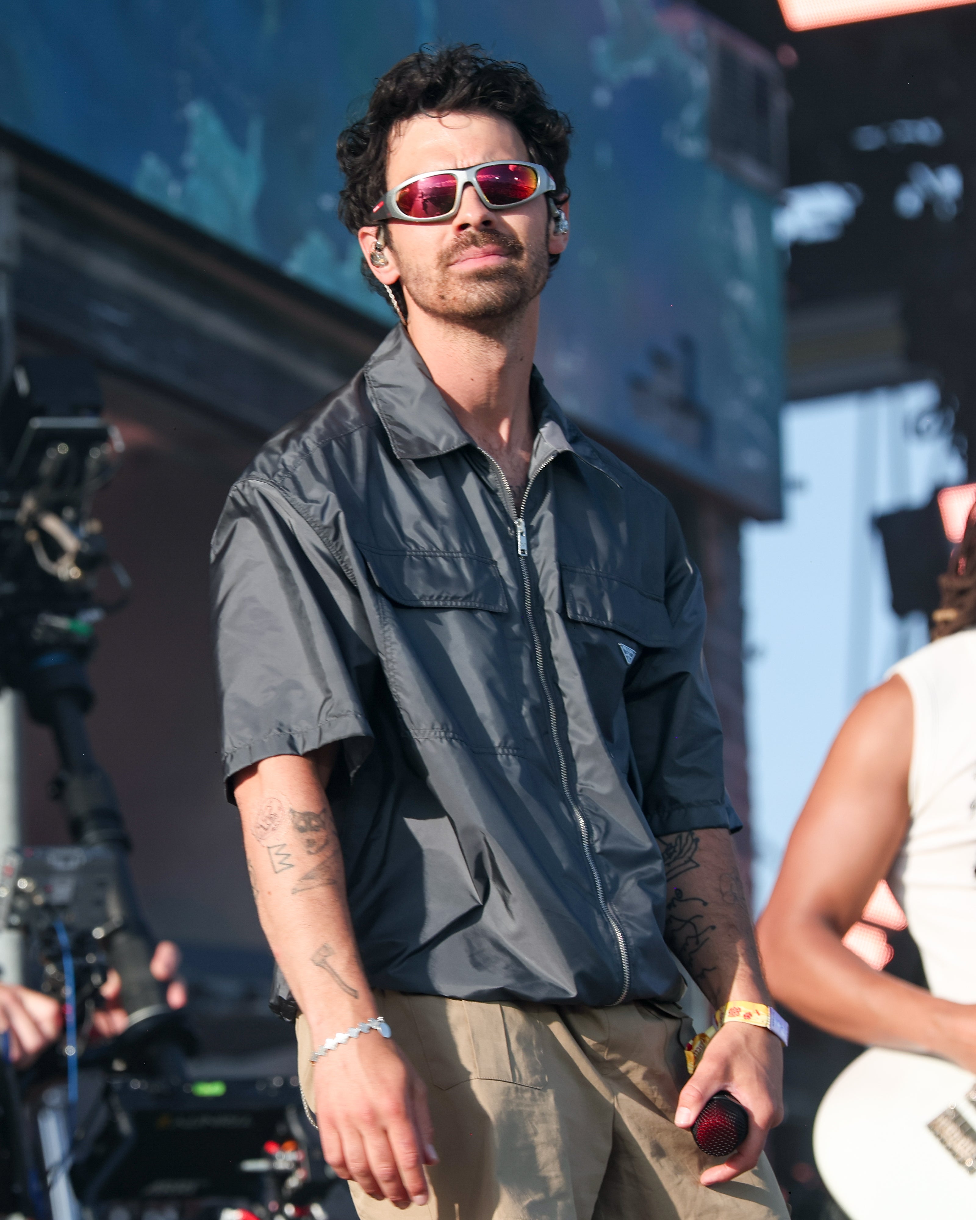Musician performing on stage wearing a casual zip-up shirt and sunglasses, holding a microphone