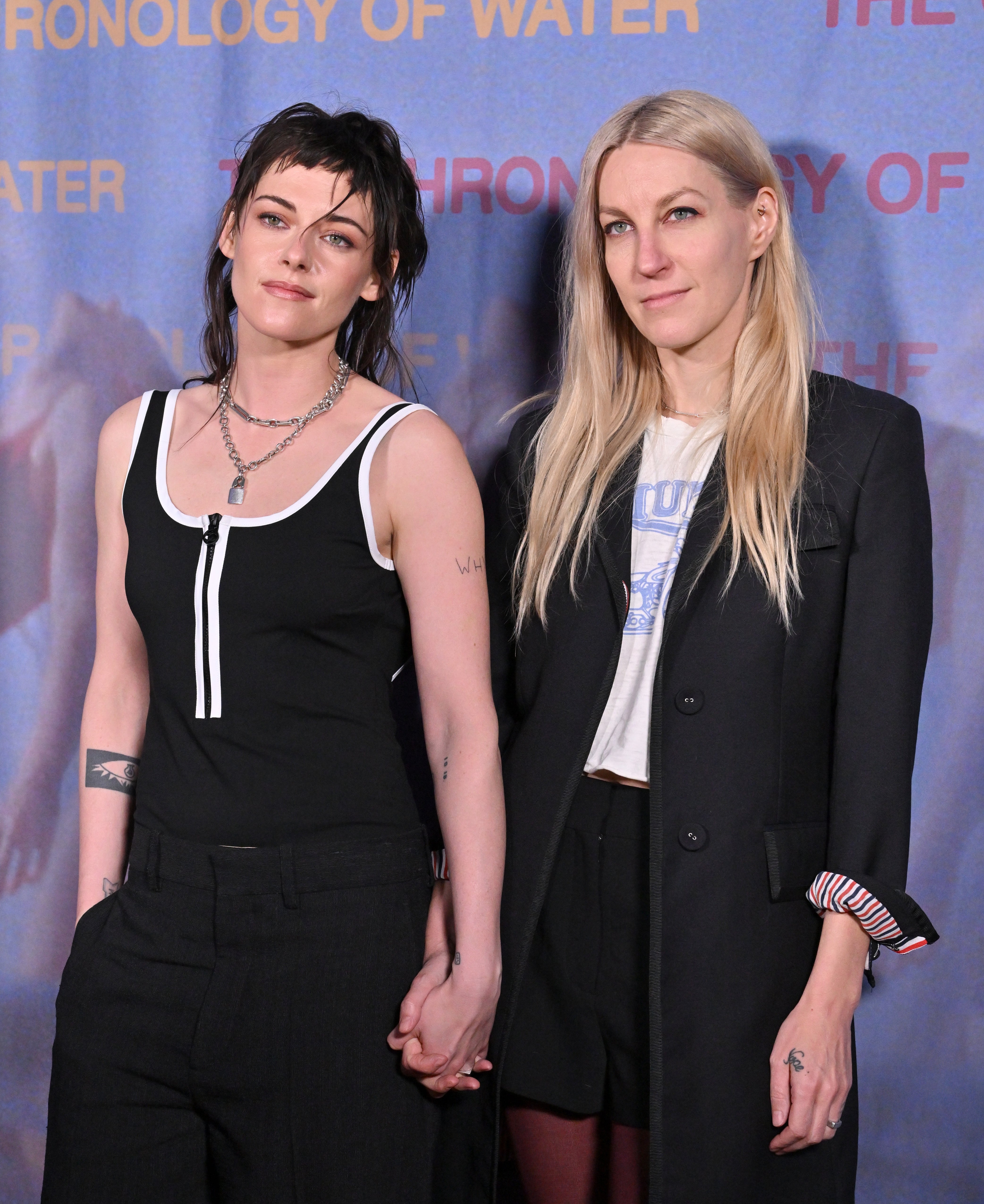 Two individuals hold hands at a red carpet event. One wears a sleeveless top and pants; the other is in a blazer with a graphic tee underneath