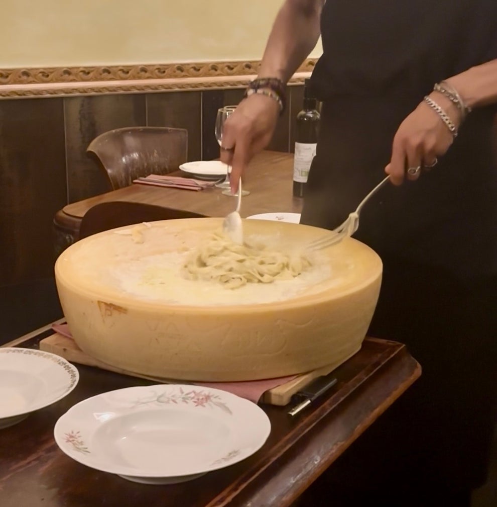 A person is serving pasta from a large wheel of cheese onto plates at a dining table, creating a unique culinary experience