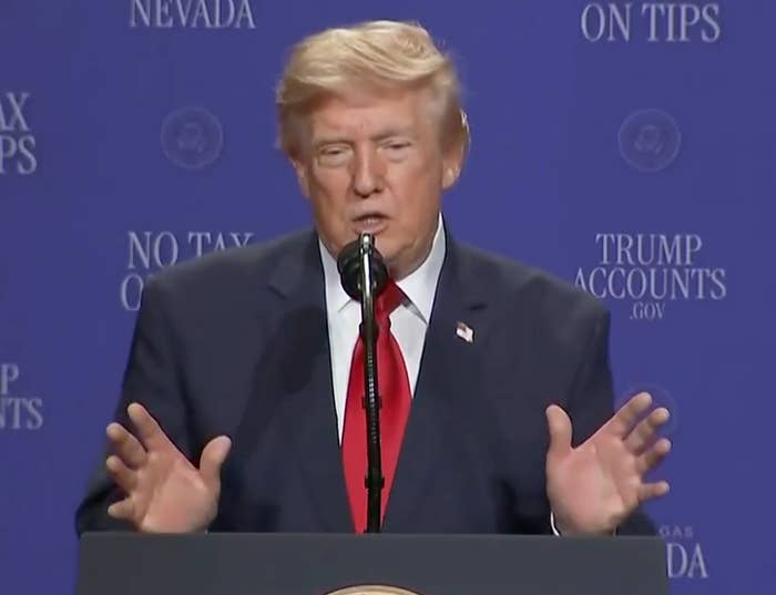 A person stands at a podium speaking, wearing a suit and red tie, in front of a backdrop with text related to tax and government topics