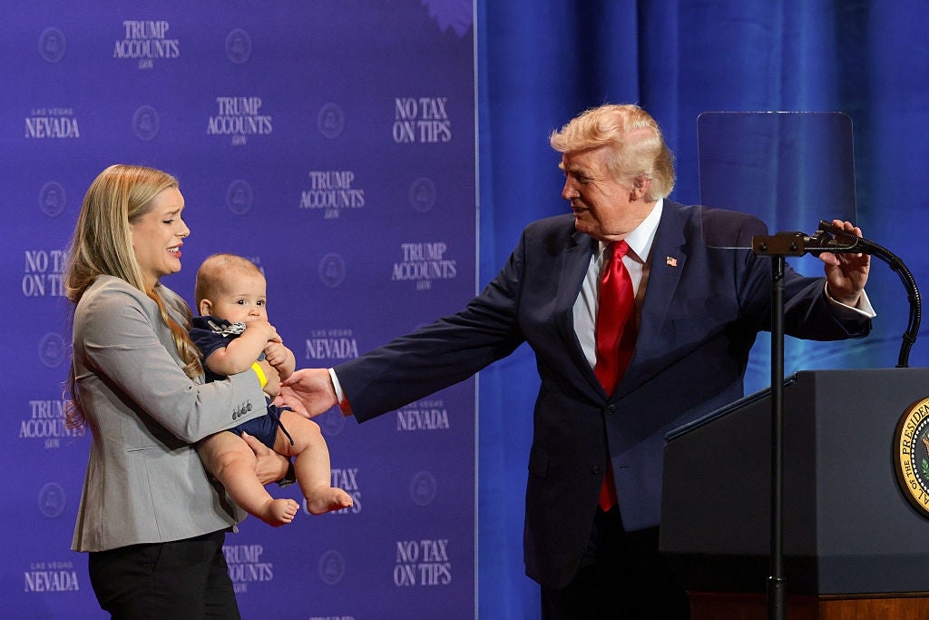 En kvinne holder en baby på scenen som en mann i dress og rødt slips beveger seg mot dem under et offentlig arrangement med en presidentsegl på podiet