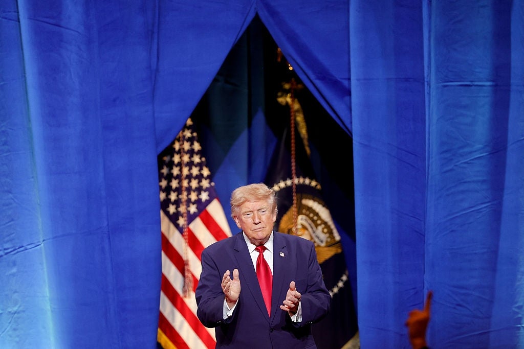 En mann i dress klapper med amerikanske flagg og en presidentsegl i bakteppet på et arrangement