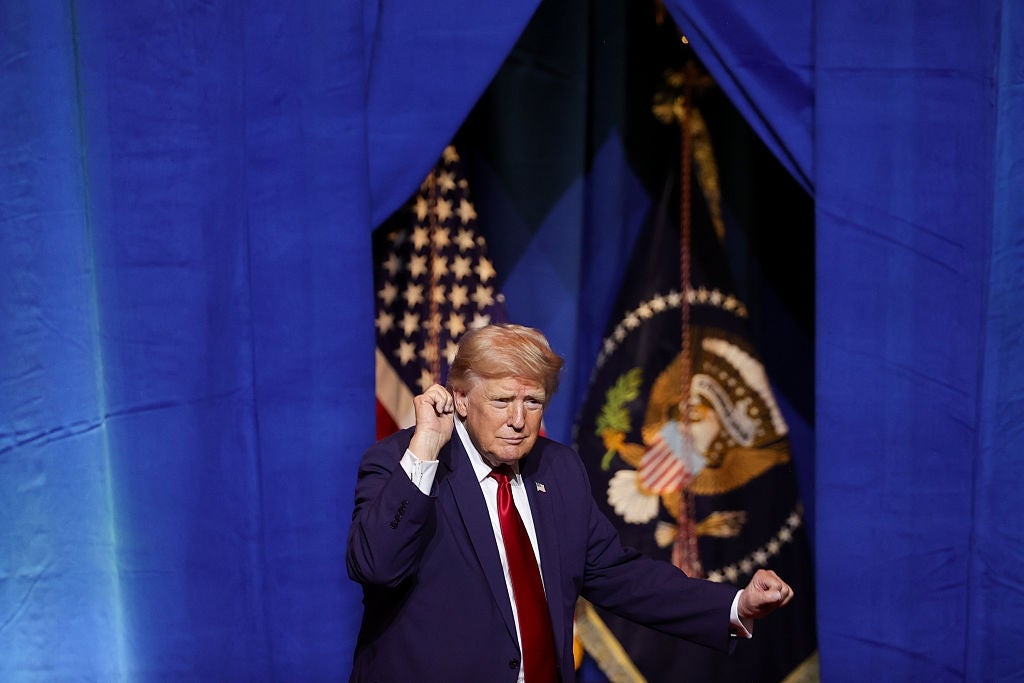 En person i dress gestikulerer mens han står foran amerikanske flagg og et presidentemblem i bakgrunnen