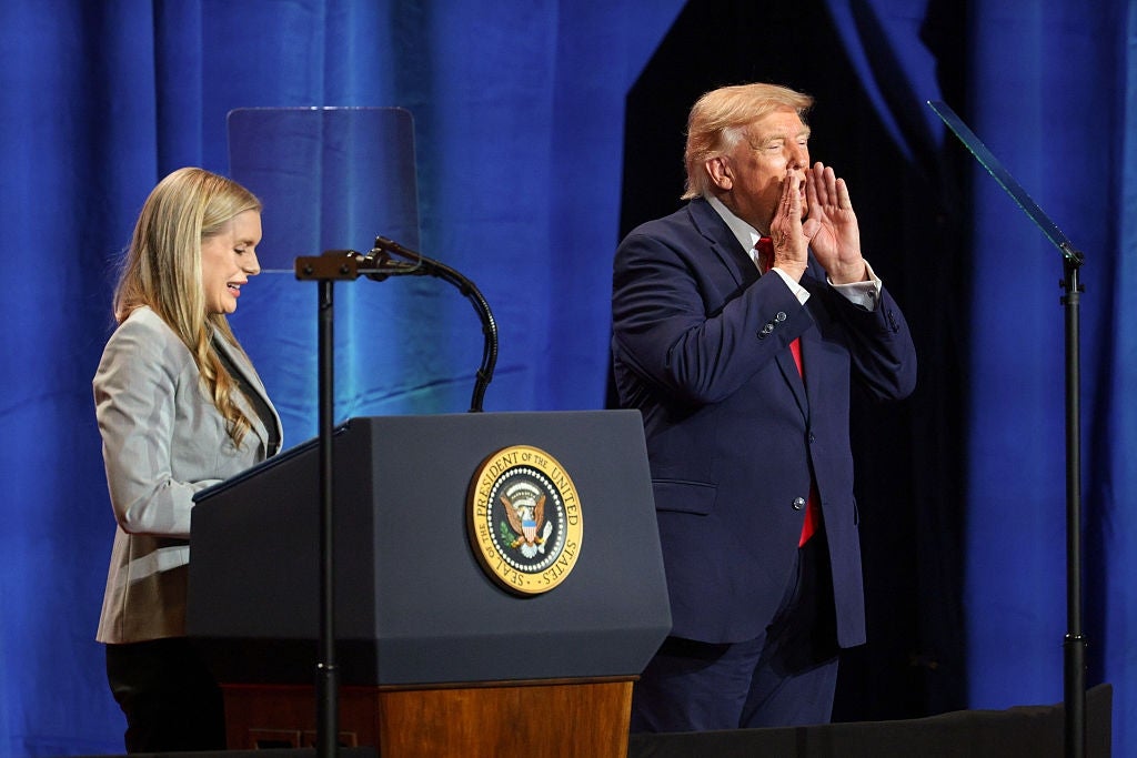 En mann i dress med en presidentsegl på scenen bevegelser, mens en kvinne står ved et podium, begge engasjert med publikum