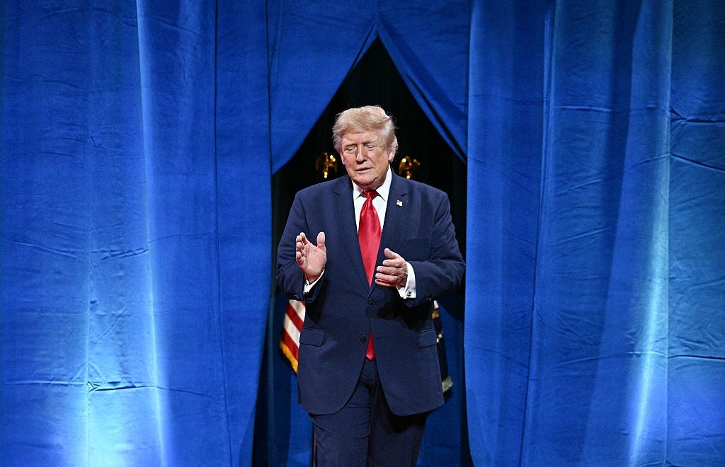 En mann i dress og rødt slips står mellom blå gardiner og gestikulerer med begge hender. Et amerikansk flagg er delvis synlig i bakgrunnen