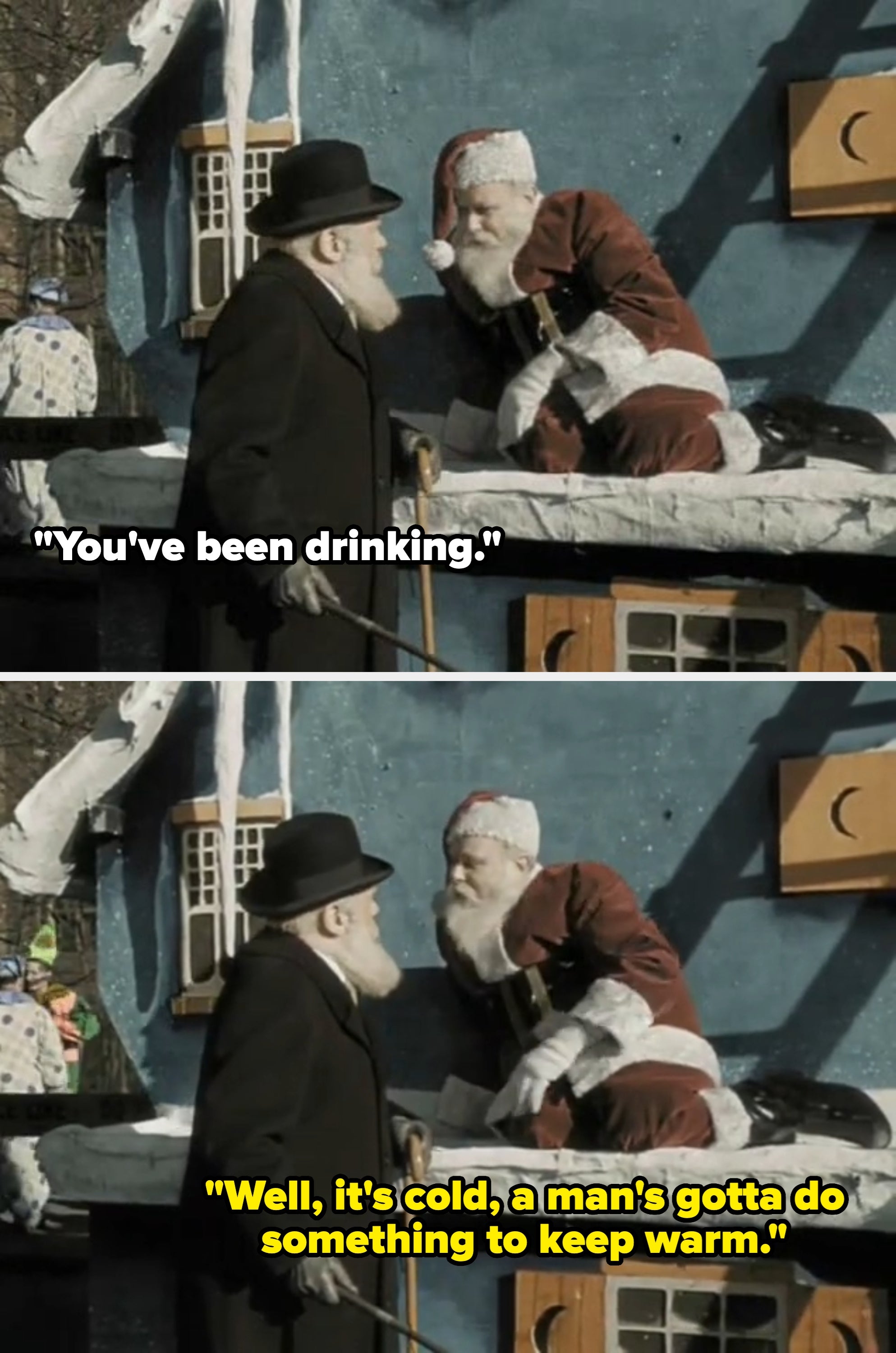 A man in a winter coat and hat talks to a person dressed as Santa Claus, sitting on a snowy roof in a holiday scene