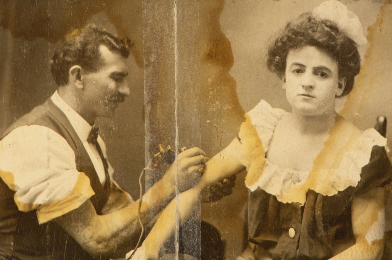 A vintage photo of a man tattooing a woman's arm. The woman wears a dress with puffed sleeves and lace, and the tattoo artist is focused on his work