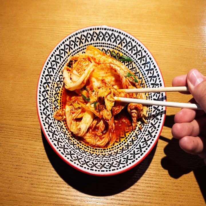 A bowl of kimchi is being picked up with chopsticks, set on a patterned plate on a wooden table