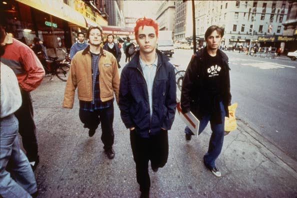 Three individuals walk down a city street. The person in the center has spiky hair and wears a casual jacket, exuding a music band vibe