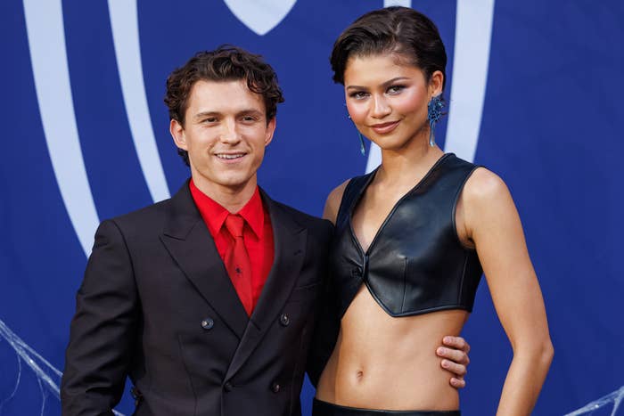Two individuals on a red carpet; one in a dark suit with a red shirt, the other in a stylish black leather crop top and trousers, posing together