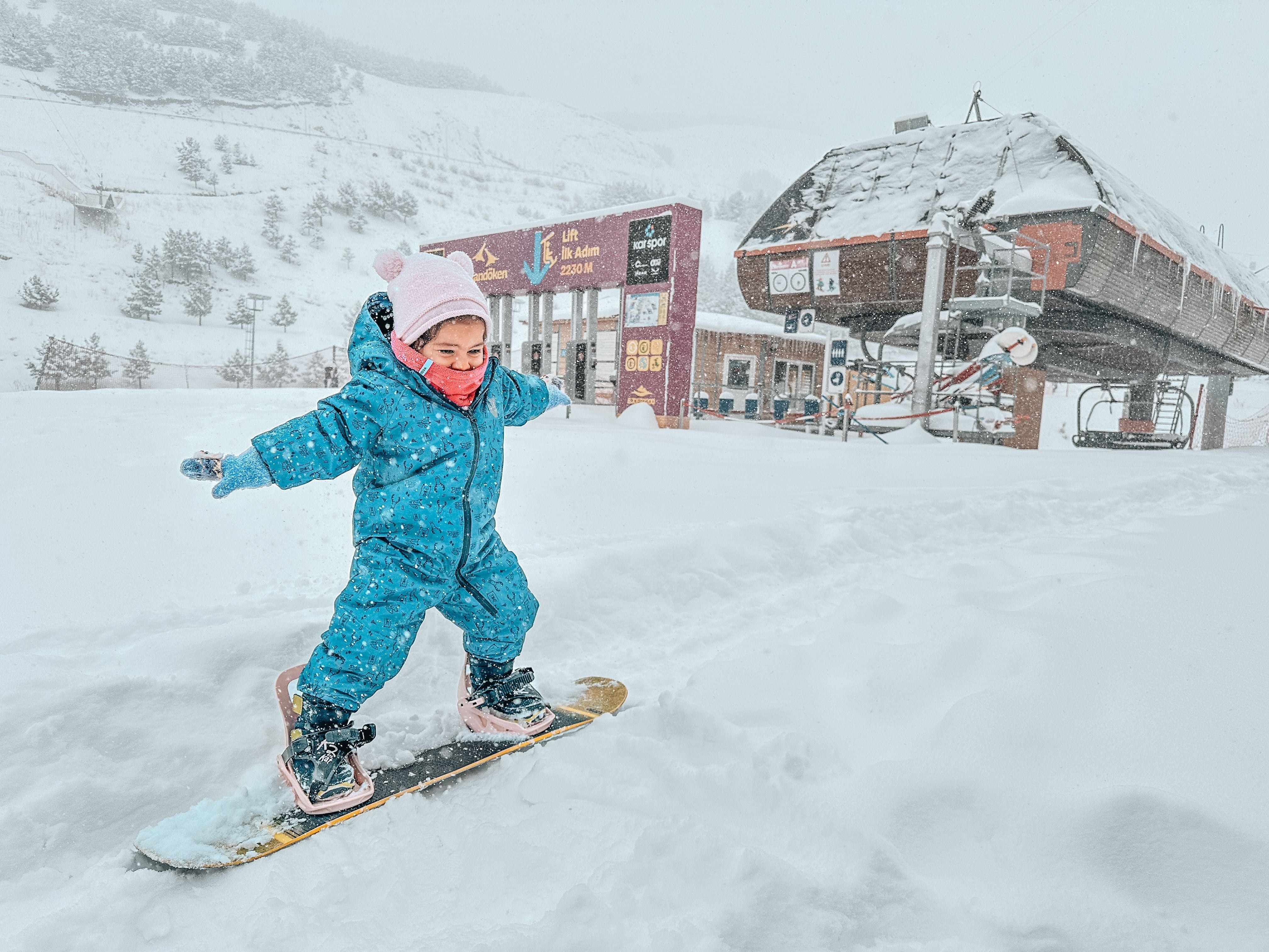 Child snowboarding in a snowsuit, hat, and mittens, joyfully navigating a snowy landscape near a ski lift and lodge