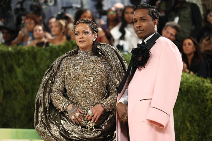 Two individuals at a formal event. One wears a shimmering, textured gown with a cape. The other sports a coat with floral accents