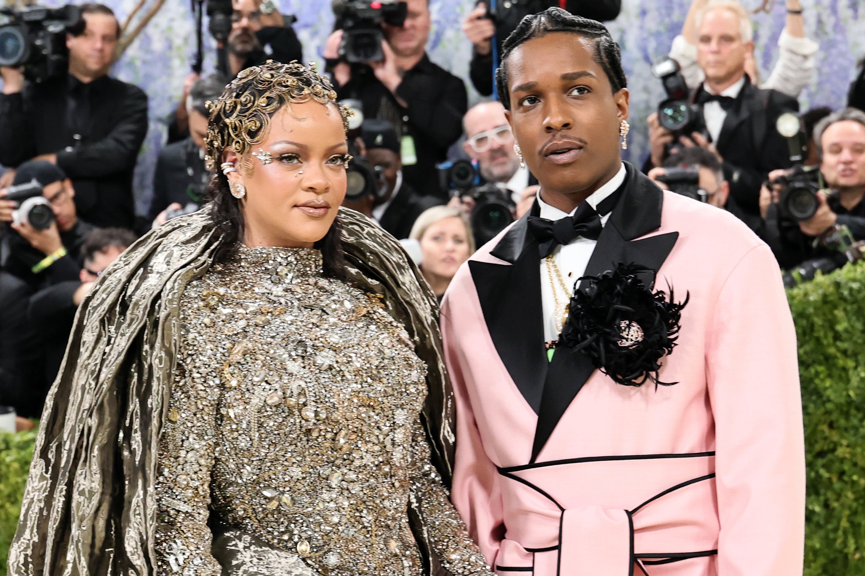 Two people on a red carpet. One wears an ornate, sequined gown with a headpiece, and the other wears a pink suit with a floral lapel detail
