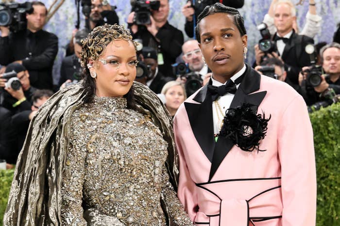 Two people on a red carpet. One wears an ornate, sequined gown with a headpiece, and the other wears a pink suit with a floral lapel detail