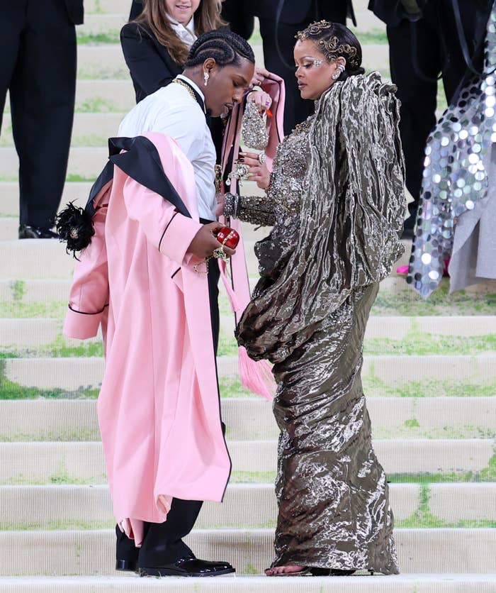 Two individuals in elaborate outfits appear on a red carpet. One wears a long ruffled dress, the other a suit with a draped pink coat