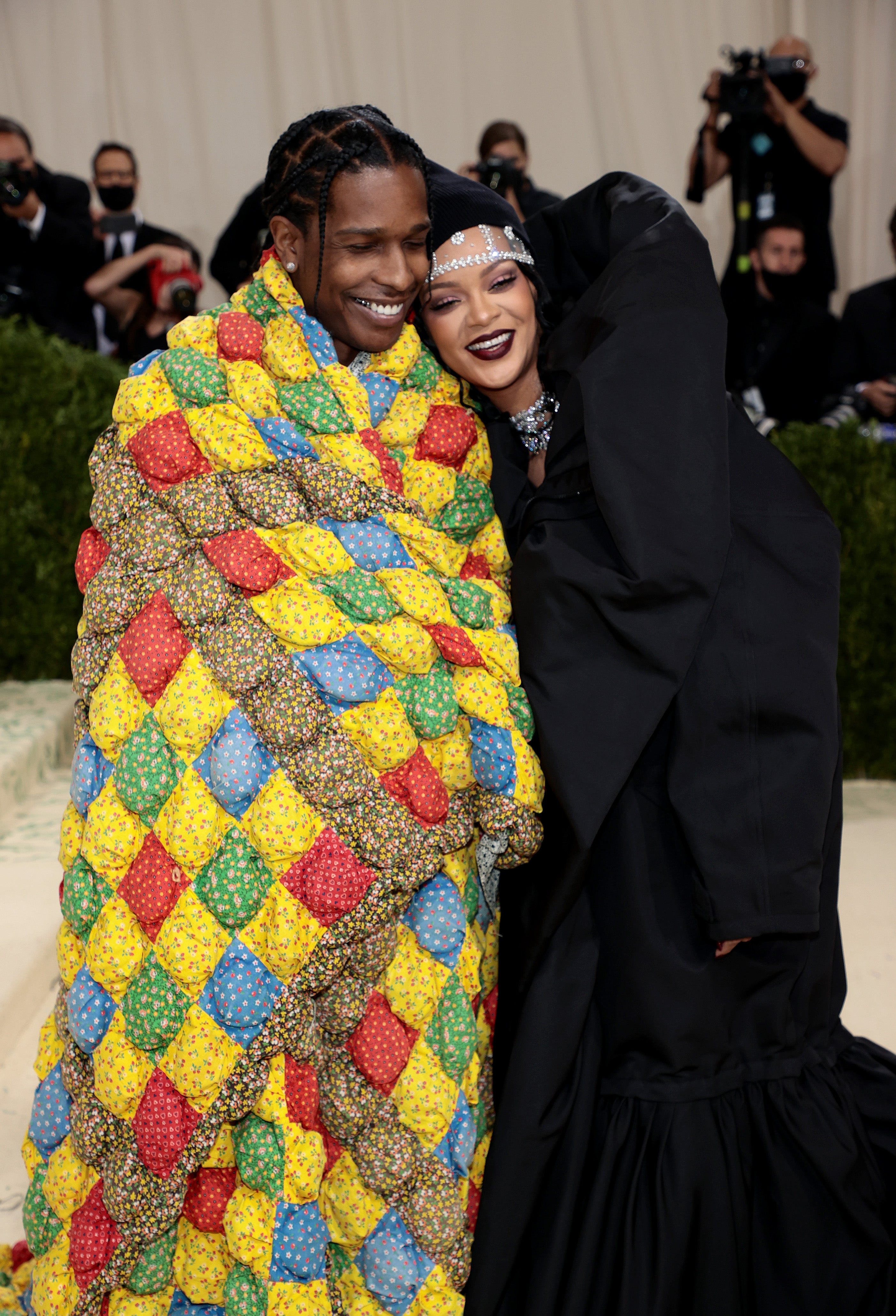 Two people smiling together on a red carpet; one in a colorful quilted blanket, the other in an elegant black outfit with a statement headpiece