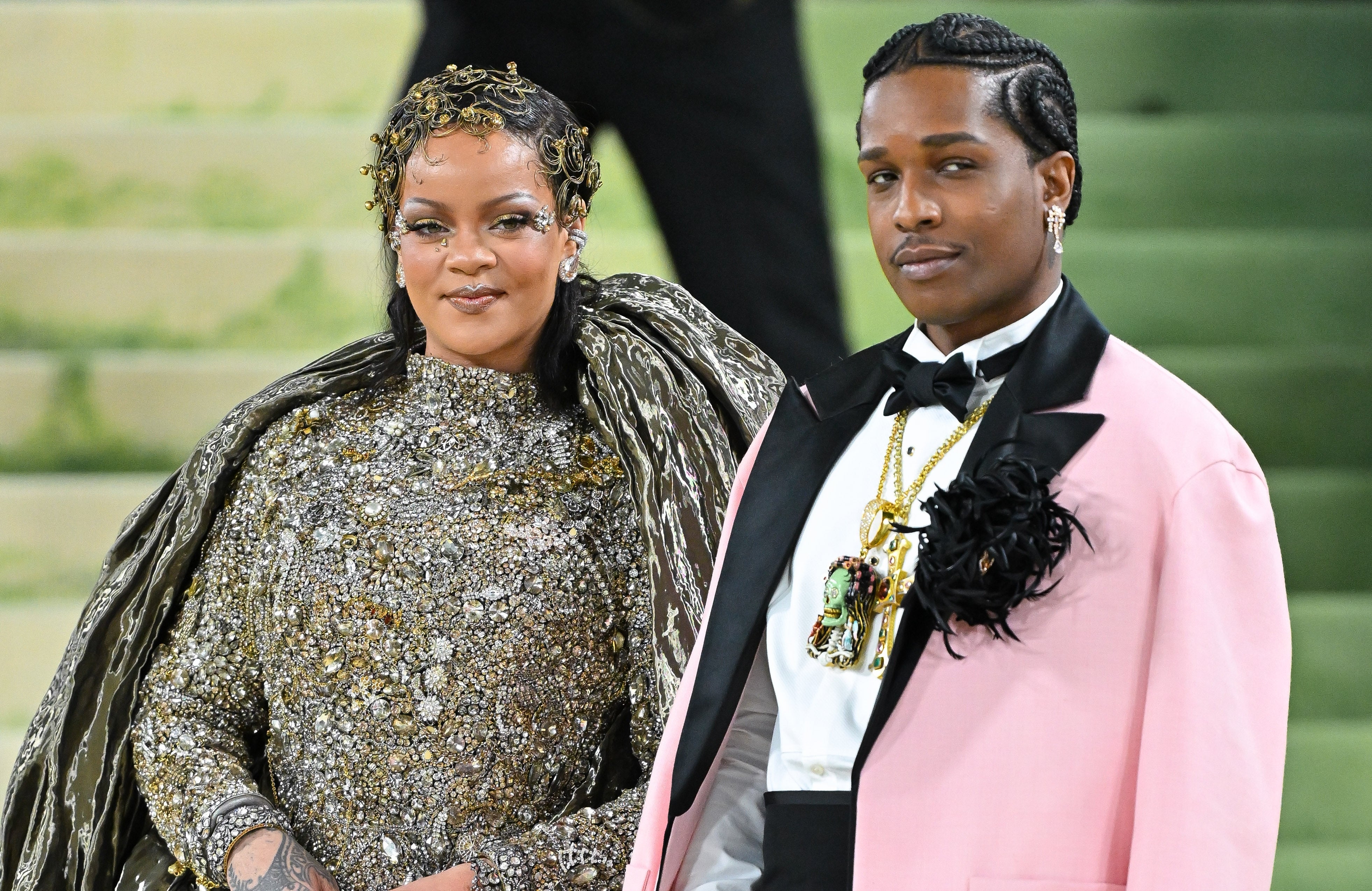 Two individuals in stylish formal attire pose on a red carpet event. The person on the left wears an ornate dress with intricate embellishments, the person on the right wears a pink jacket and bow tie