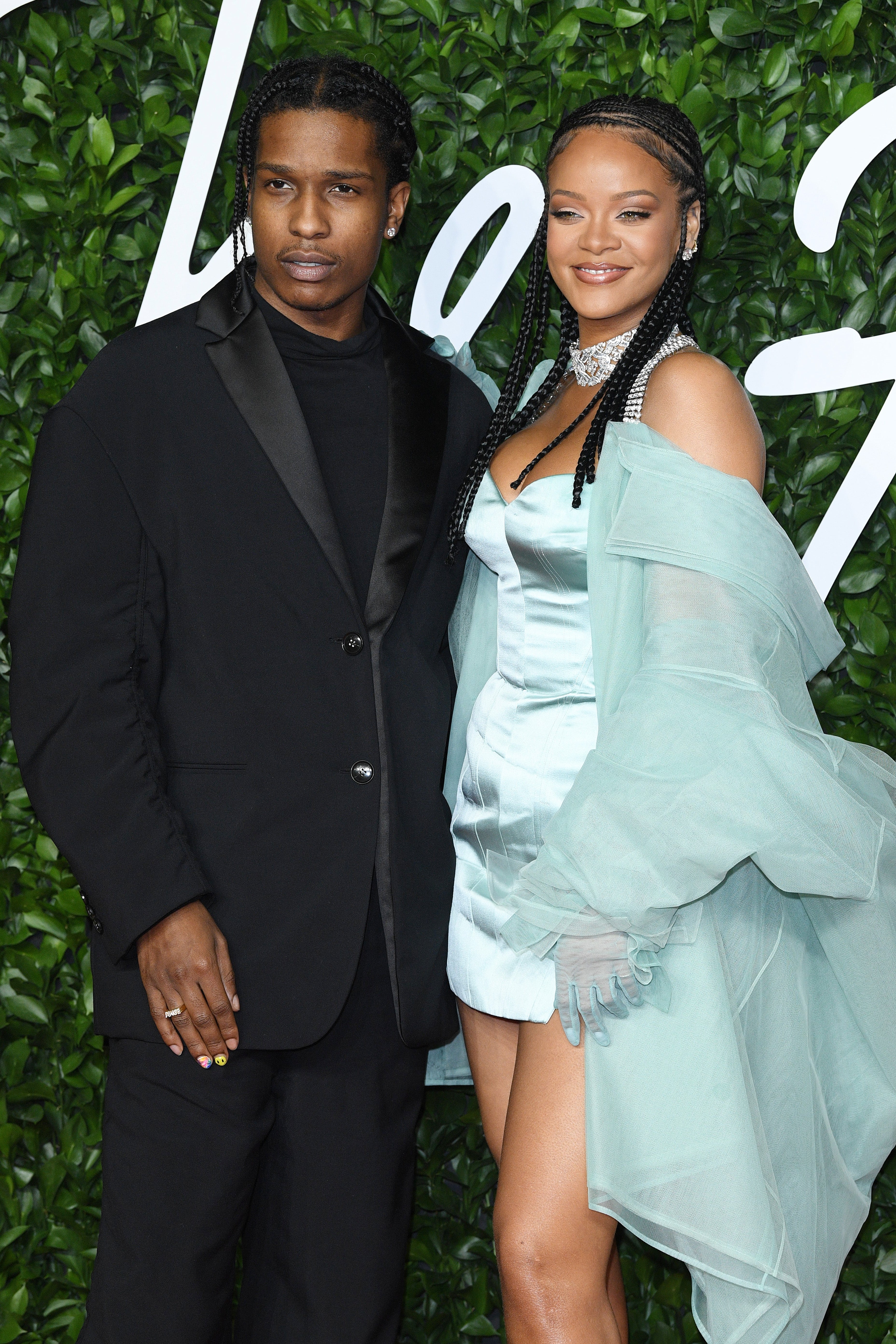 Two people posing on a green-leafed backdrop. One wears a black suit; the other in a silky dress and sheer wrap with braids