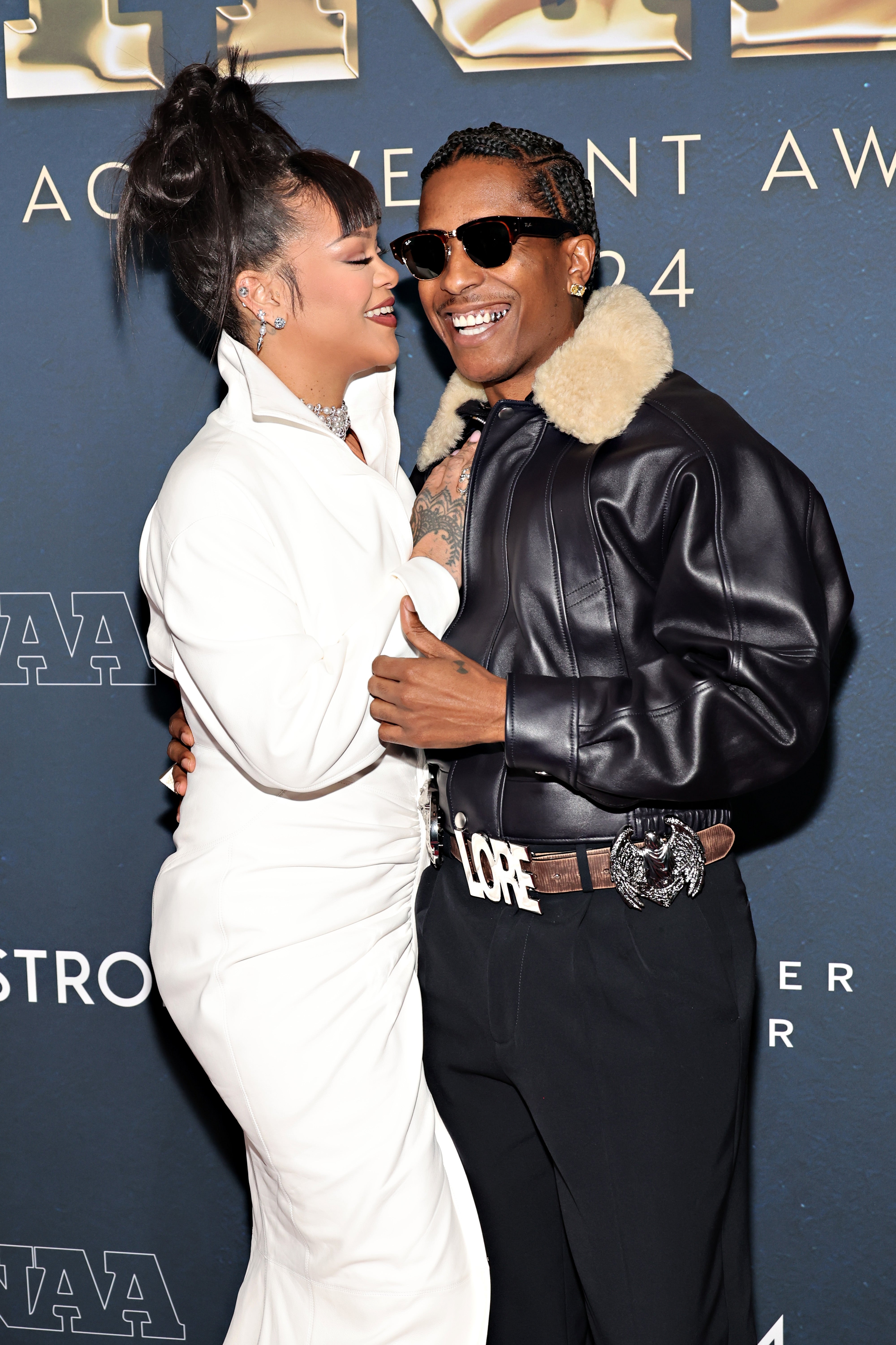 Two celebrities embrace, one in a white suit and the other in a leather jacket with a shearling collar, smiling on a red carpet