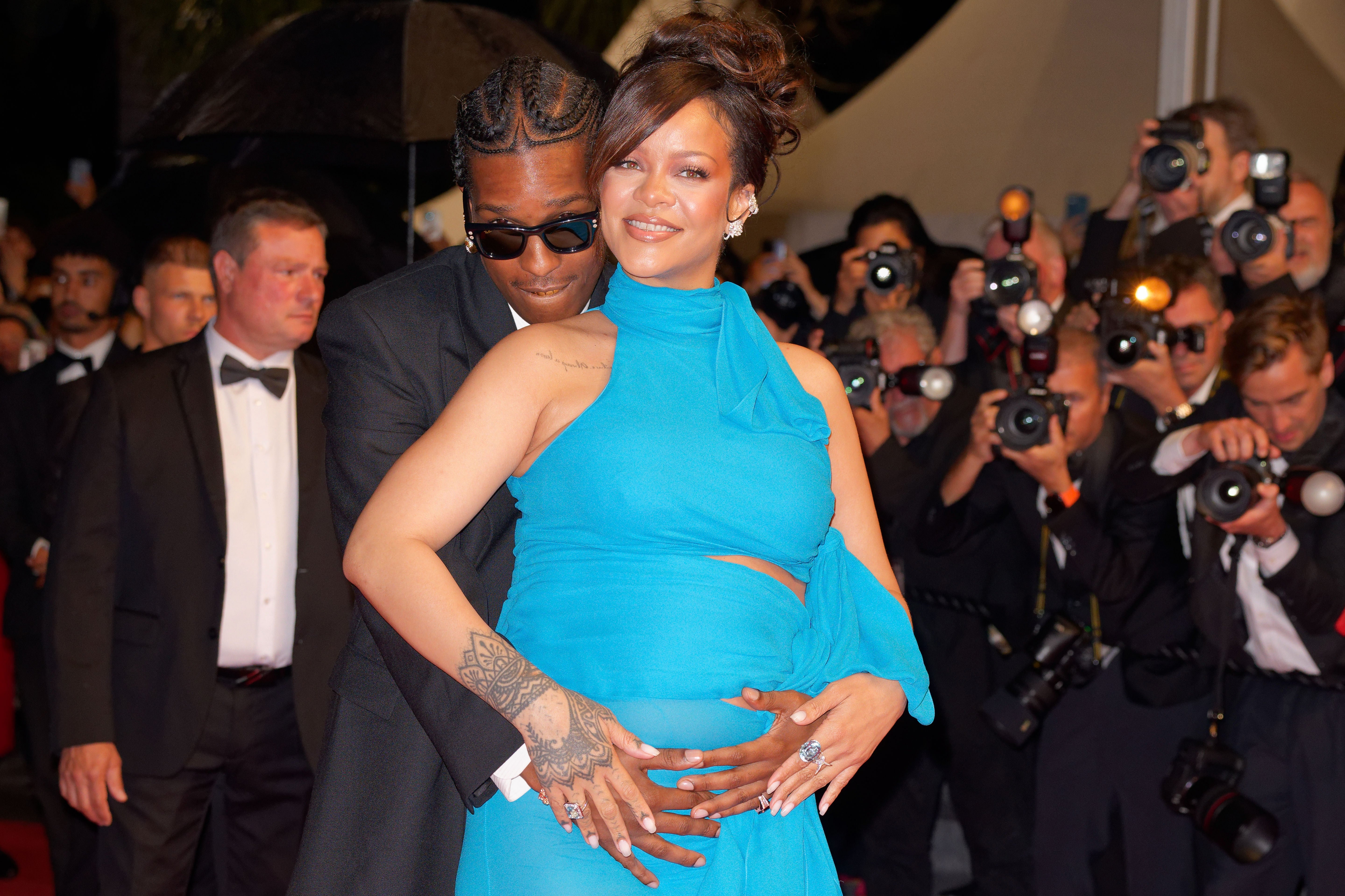 Two people pose on a red carpet. One wears a blue gown with a unique neckline, while the other is in a dark suit and sunglasses. Photographers are in the background