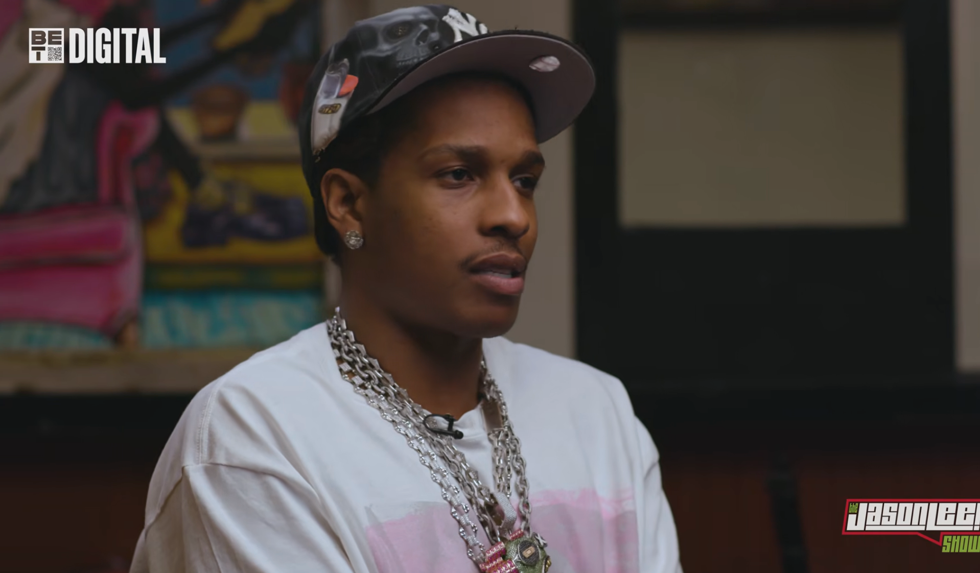A person wearing a cap, white T-shirt, and multiple silver chains sits for an interview on a digital show